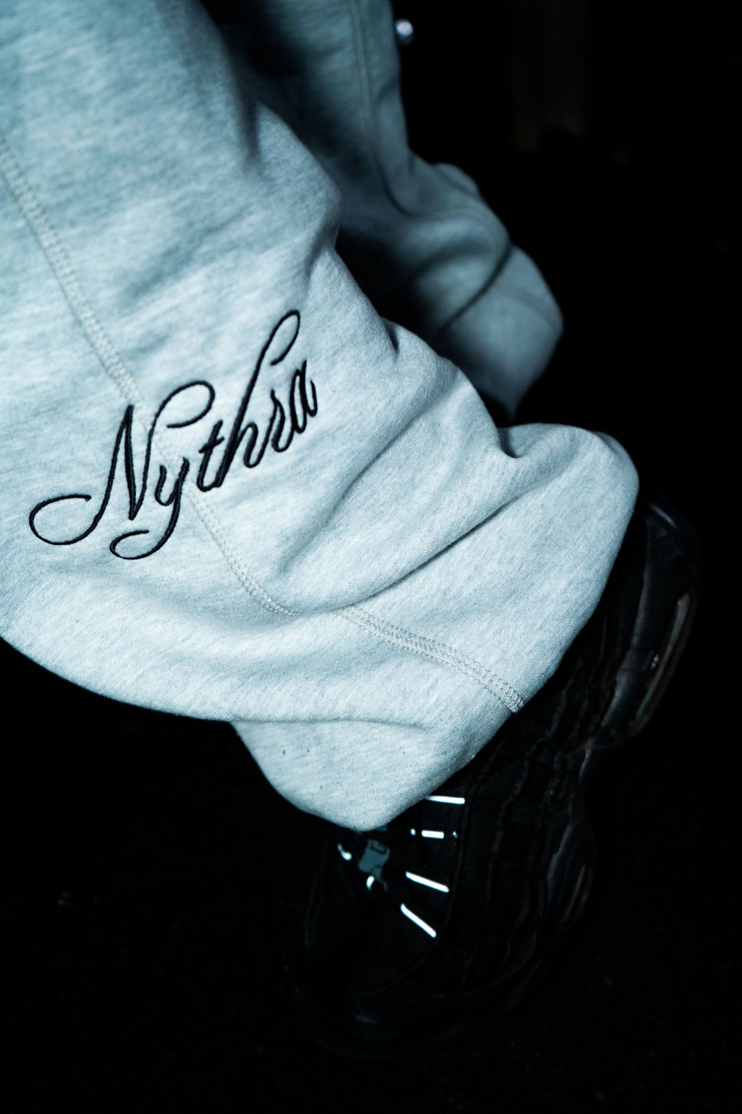 Close-up of grey sweatshirt cuff with Nythro embroidered logo and black sneaker visible.
