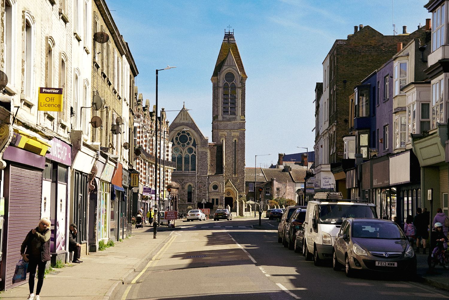 A bustling UK high street lined with shops leads to a Gothic church under a clear blue sky.