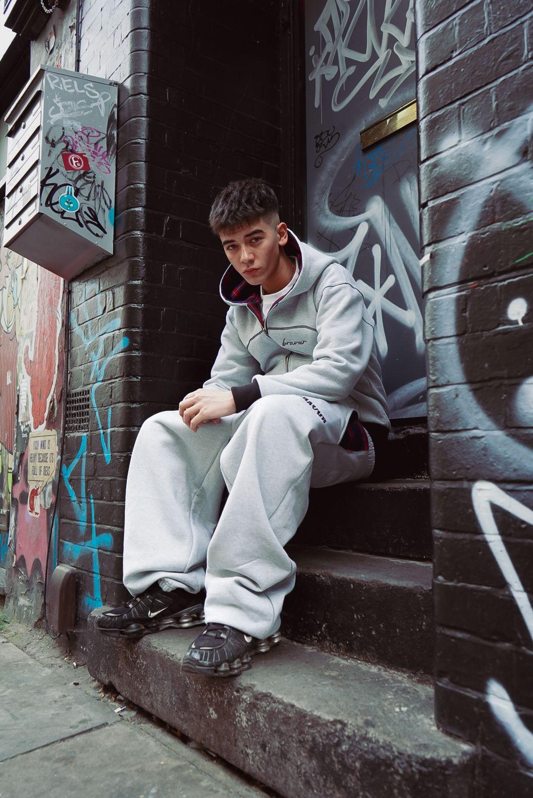 Young man in grey Bravur tracksuit sits on steps in a graffiti-covered urban alleyway.