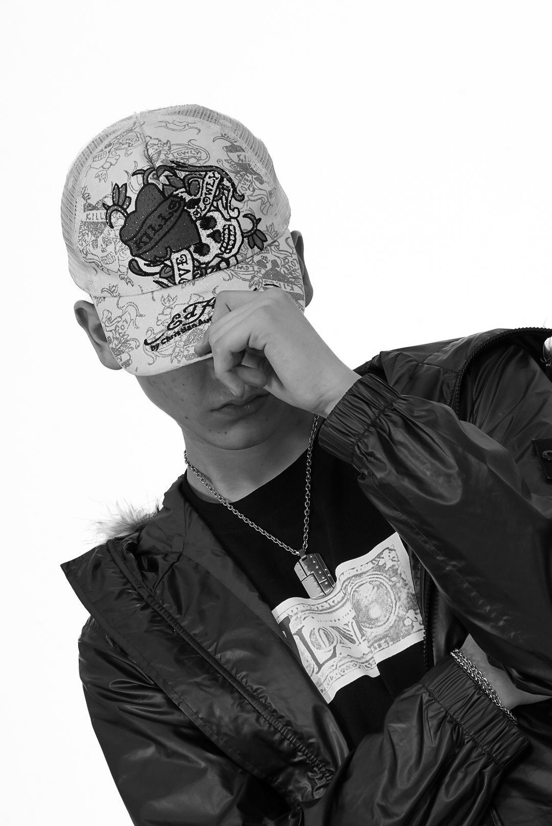 Black and white photo of young man in bomber jacket tilting cap over face with chain necklace.