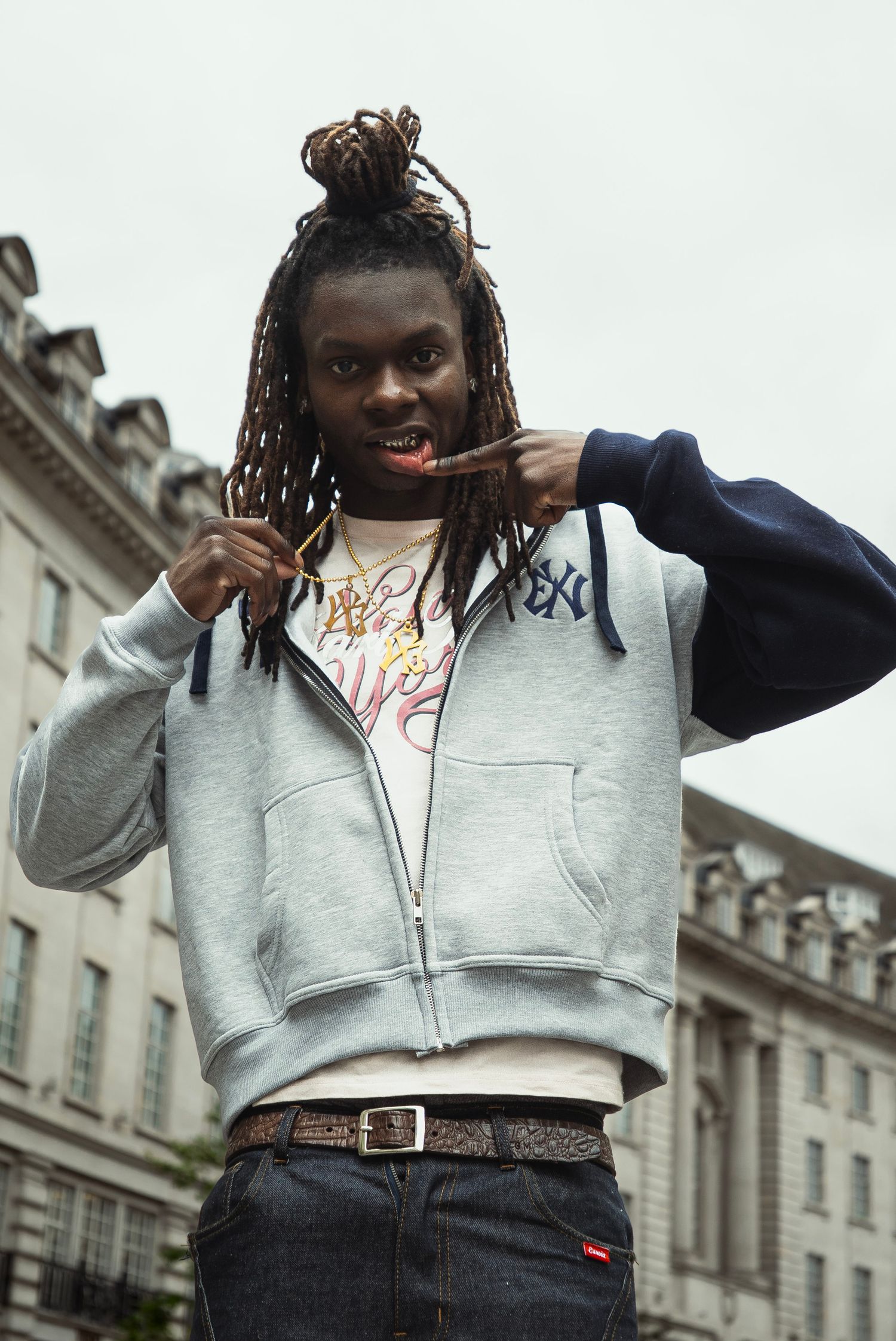 Young Black man with dreadlocks wearing a grey NY Yankees hoodie poses confidently on a city street.