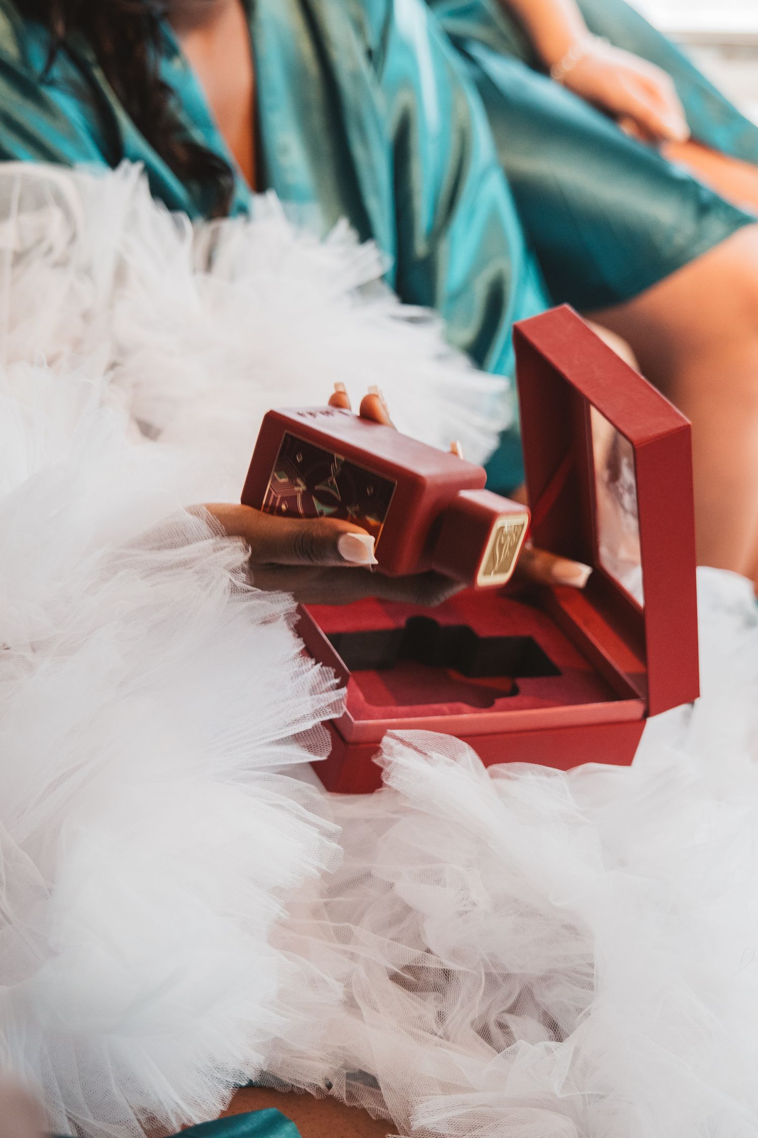 Red jewelry box nestled in white tulle fabric with teal-robed figures in background.