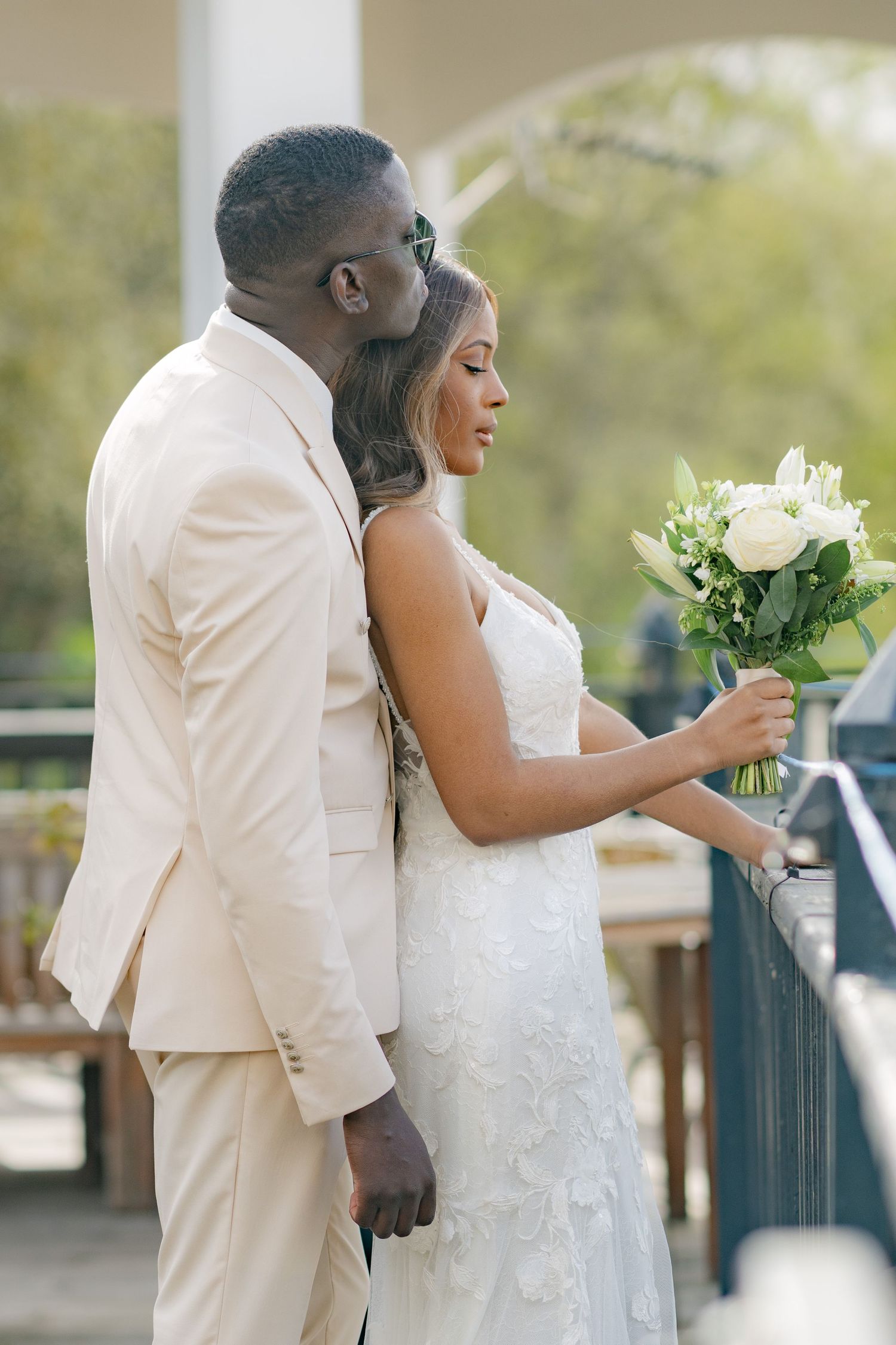 A couple in white wedding attire shares an intimate moment on a terrace with white flowers.