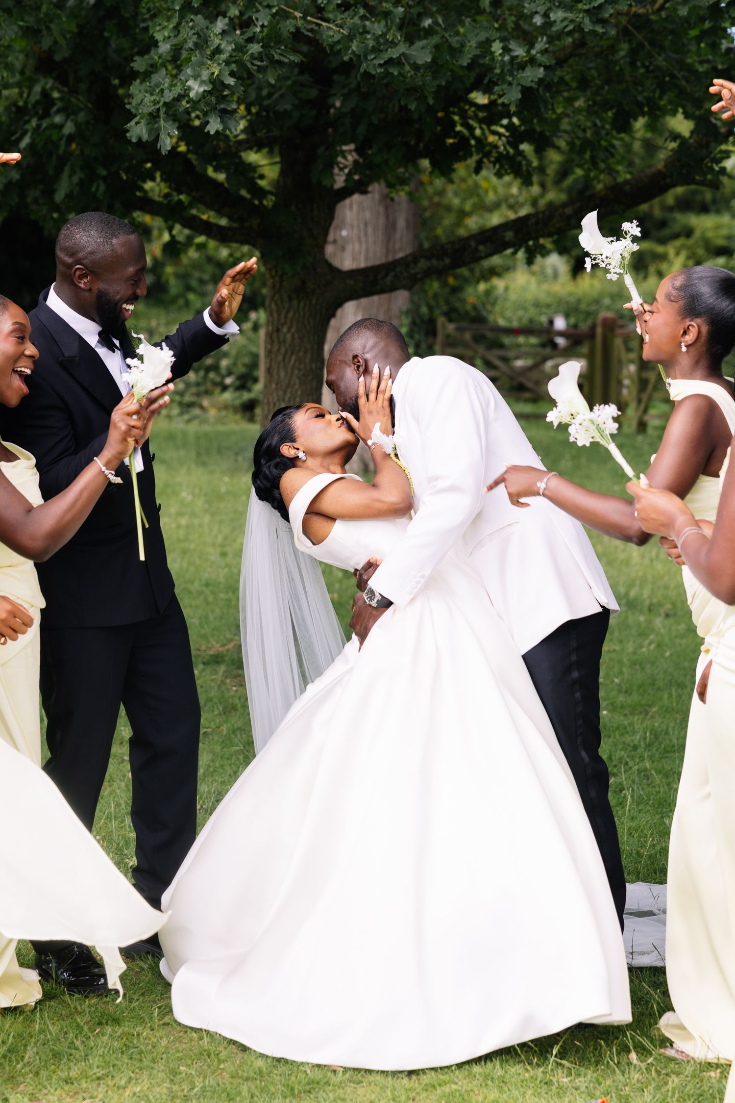Bride and groom share a romantic kiss surrounded by bridesmaids in yellow dresses and groomsmen during outdoor wedding celebration.