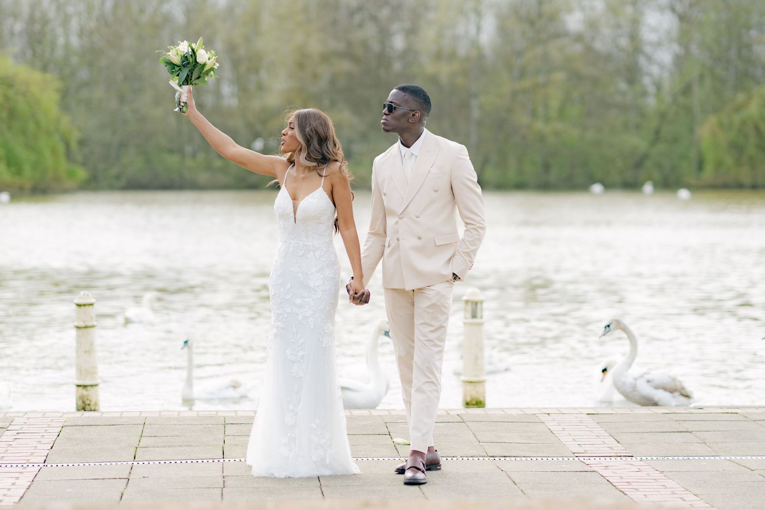 Newlyweds pose together on a stone patio overlooking a lake with swans.