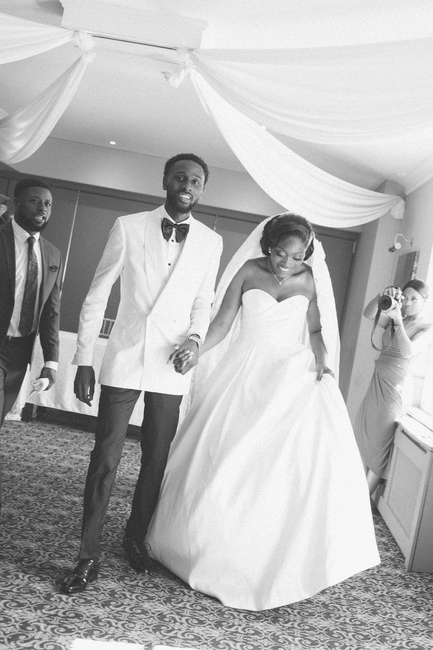 Wedding party members walking through an elegant white-draped venue hallway.