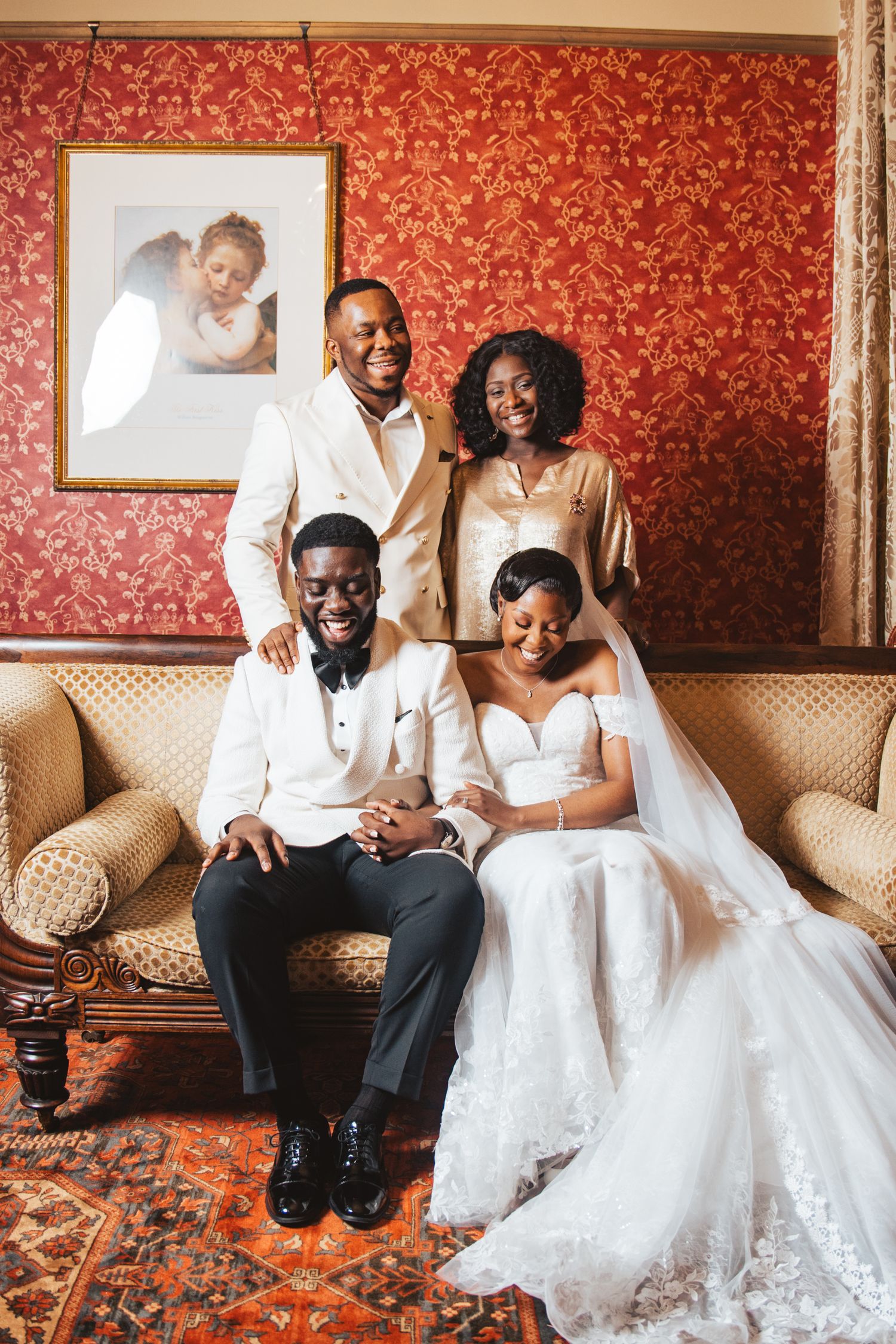 Newlyweds share intimate moments on an elegant couch against red wallpaper.