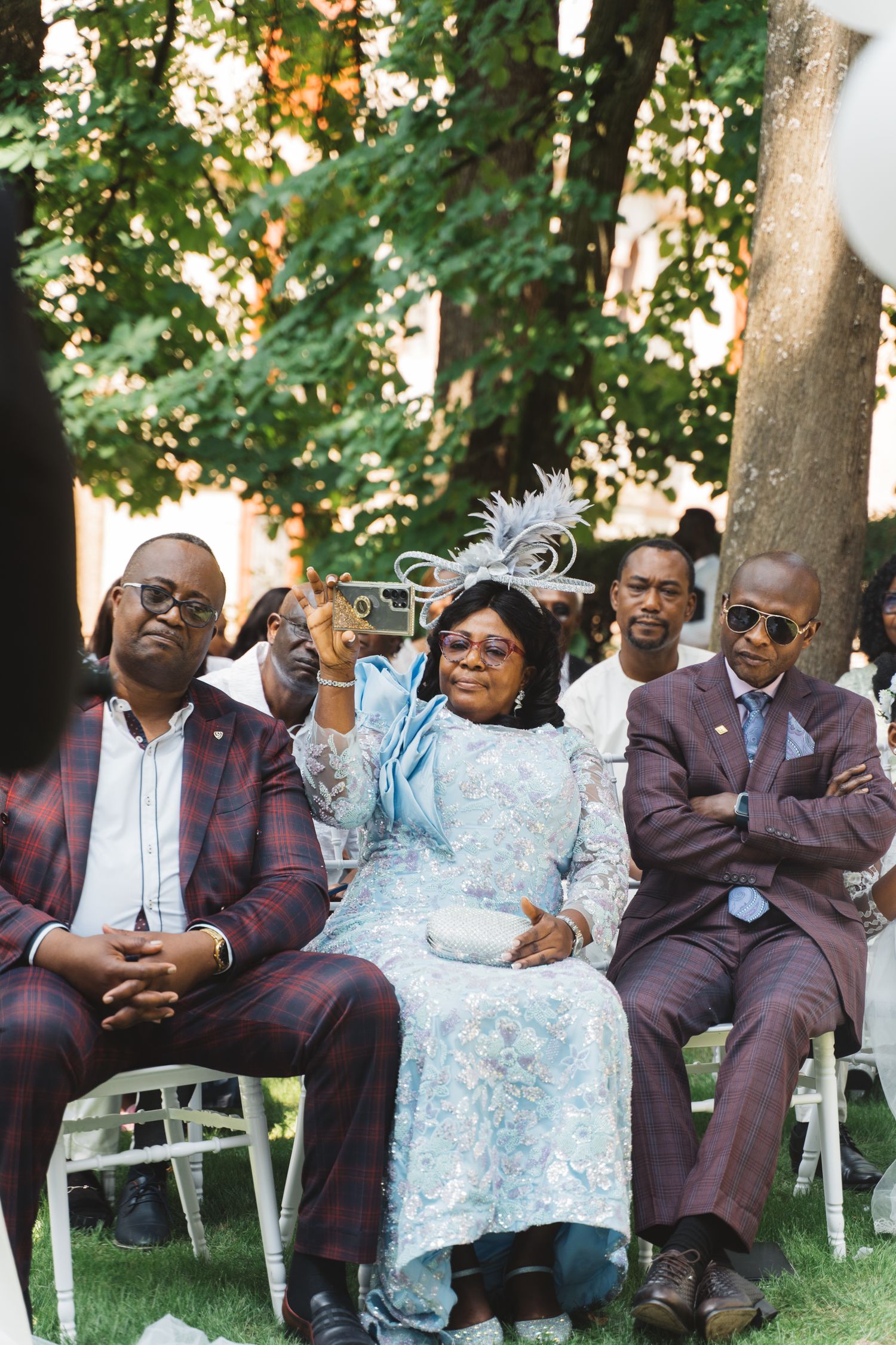 Wedding attendees in formal attire seated in white chairs during an outdoor ceremony.