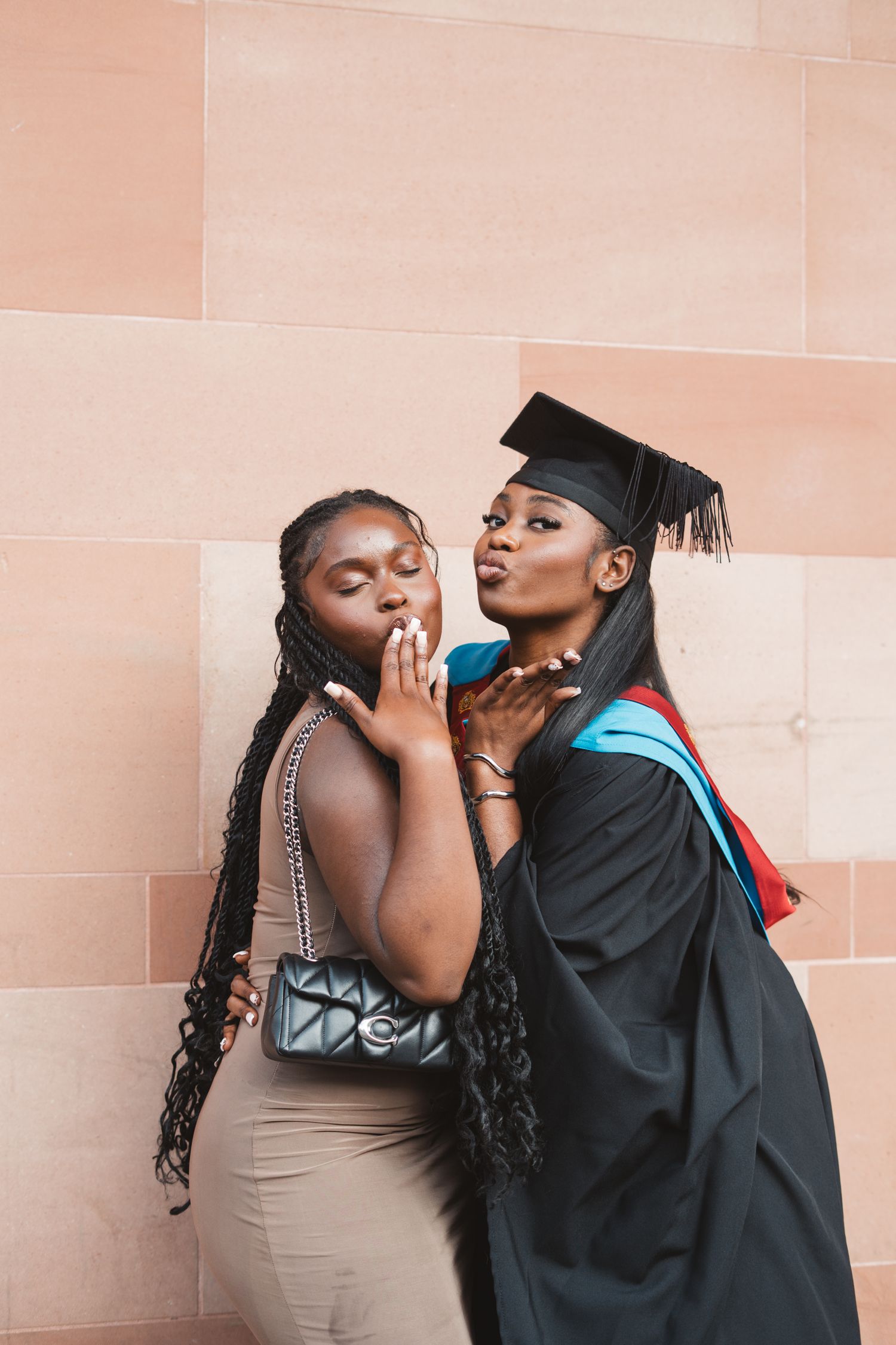 Two friends pose playfully against a pink wall during graduation celebrations.