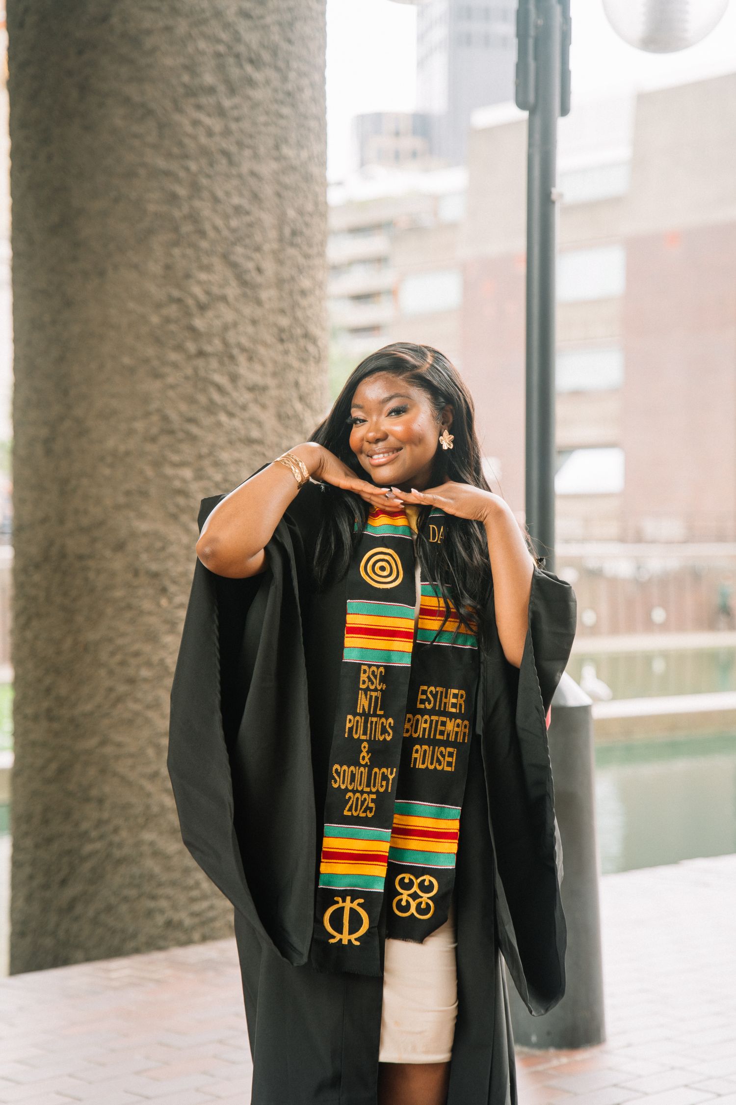 Graduate wearing academic regalia and cultural stole poses confidently under architectural columns.