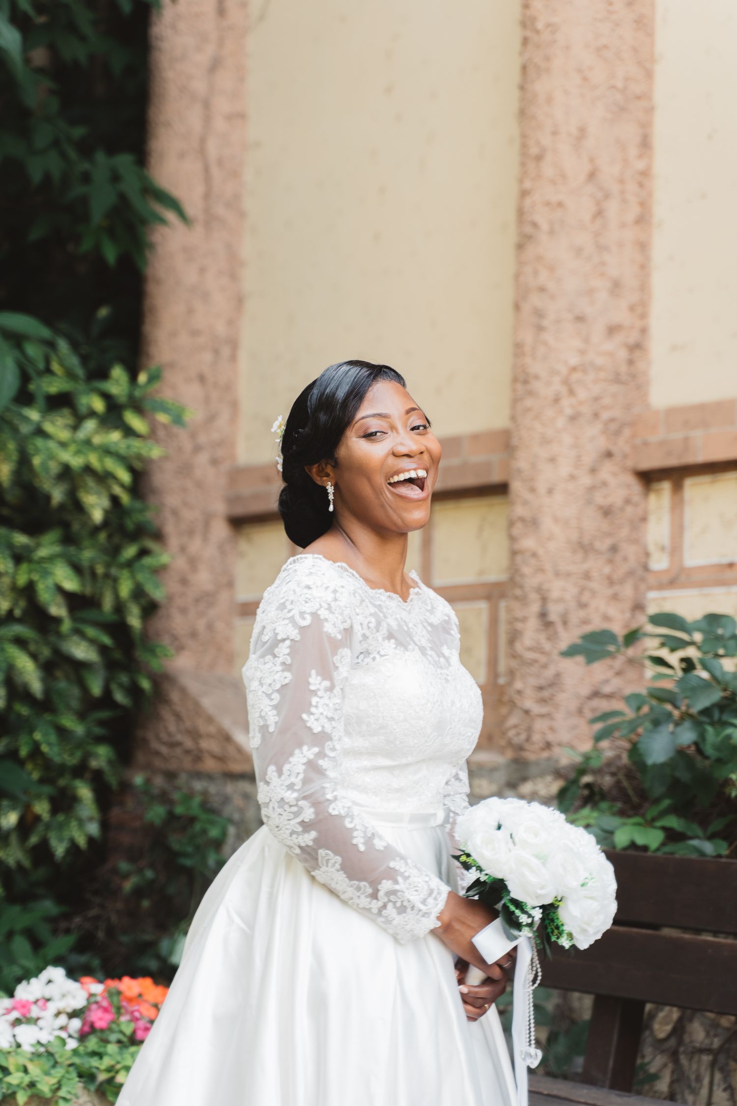 Bride in elegant lace-sleeved wedding gown sitting on bench with white bouquet.