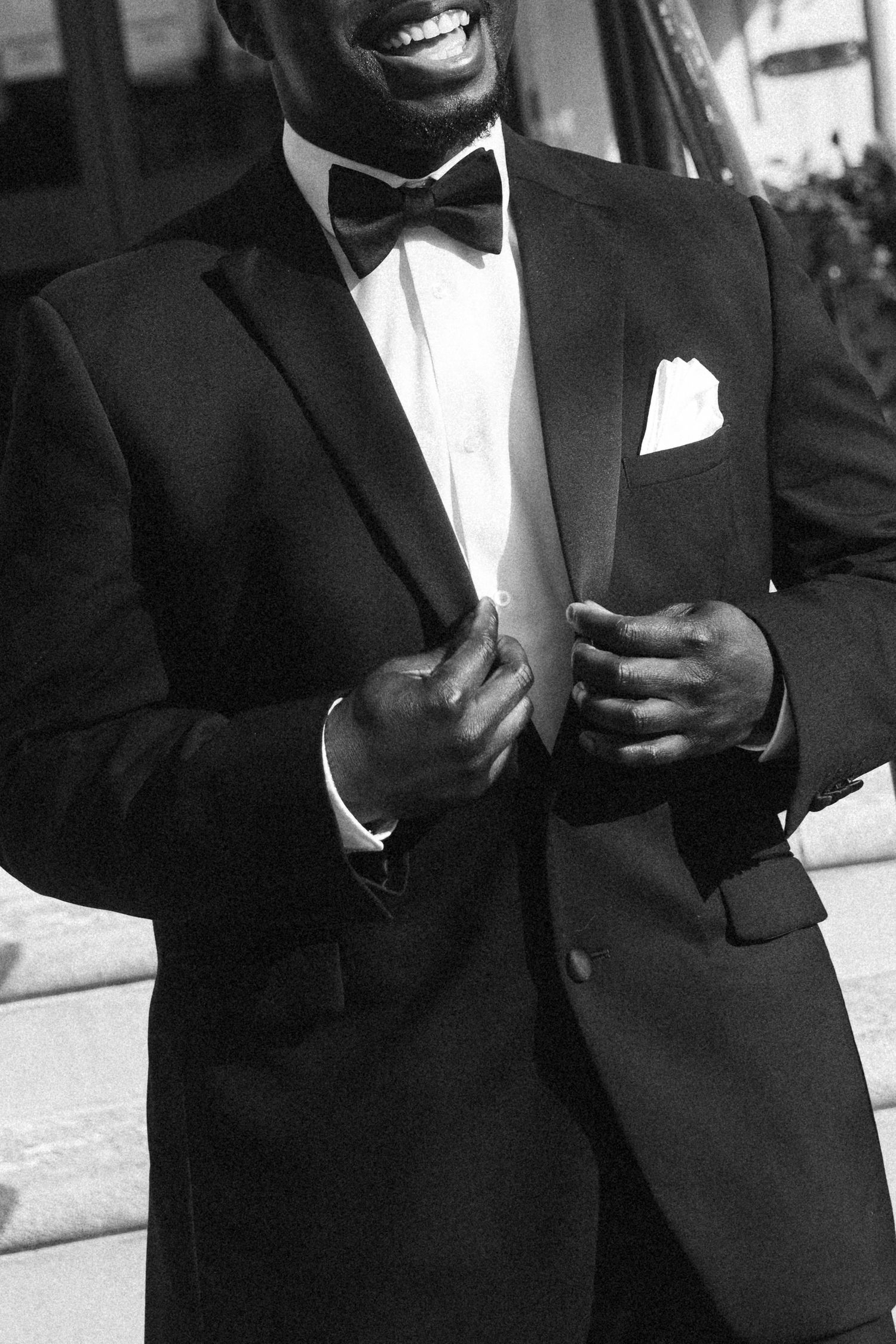 Smiling man in black tuxedo with bow tie and white pocket square adjusting his jacket lapels.