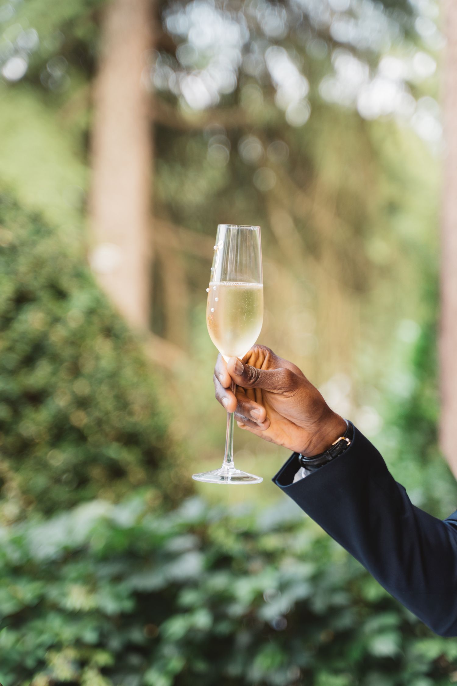 A hand holding up a champagne flute against a lush green garden background.