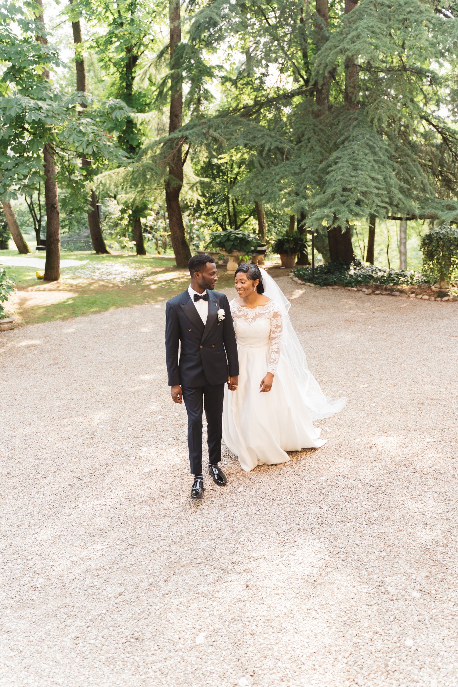 Couple walking hand in hand along a tree-lined garden path in wedding attire.