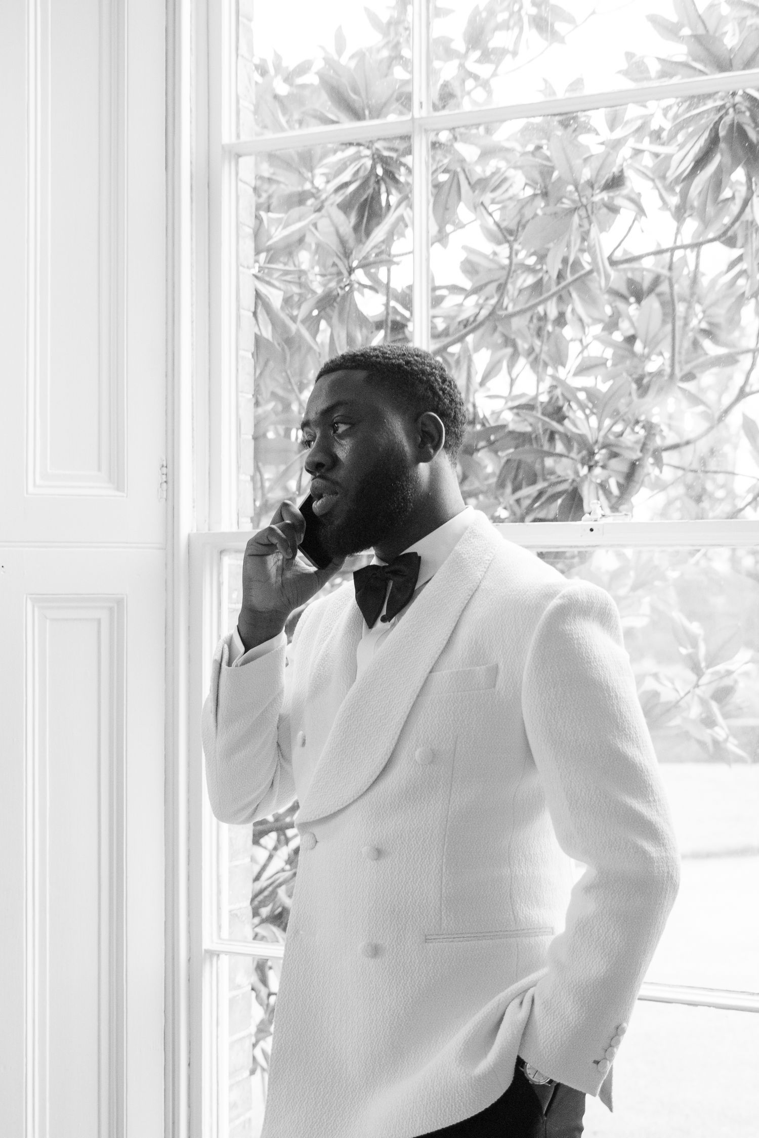 A person in a crisp white suit stands contemplatively by a bright window.