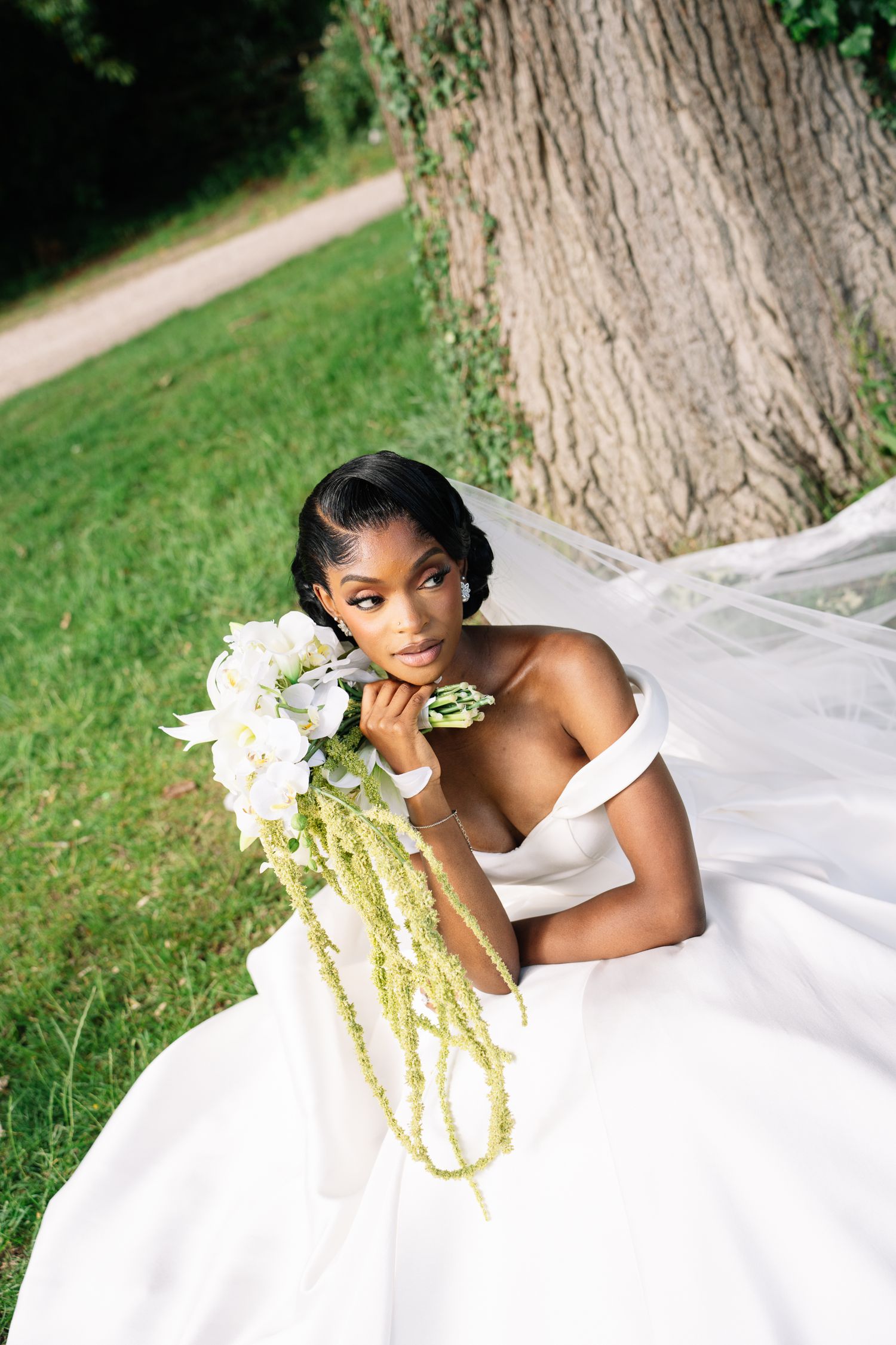 Bride in white off-shoulder gown sits by tree holding cascading bouquet of white flowers and green amaranthus.