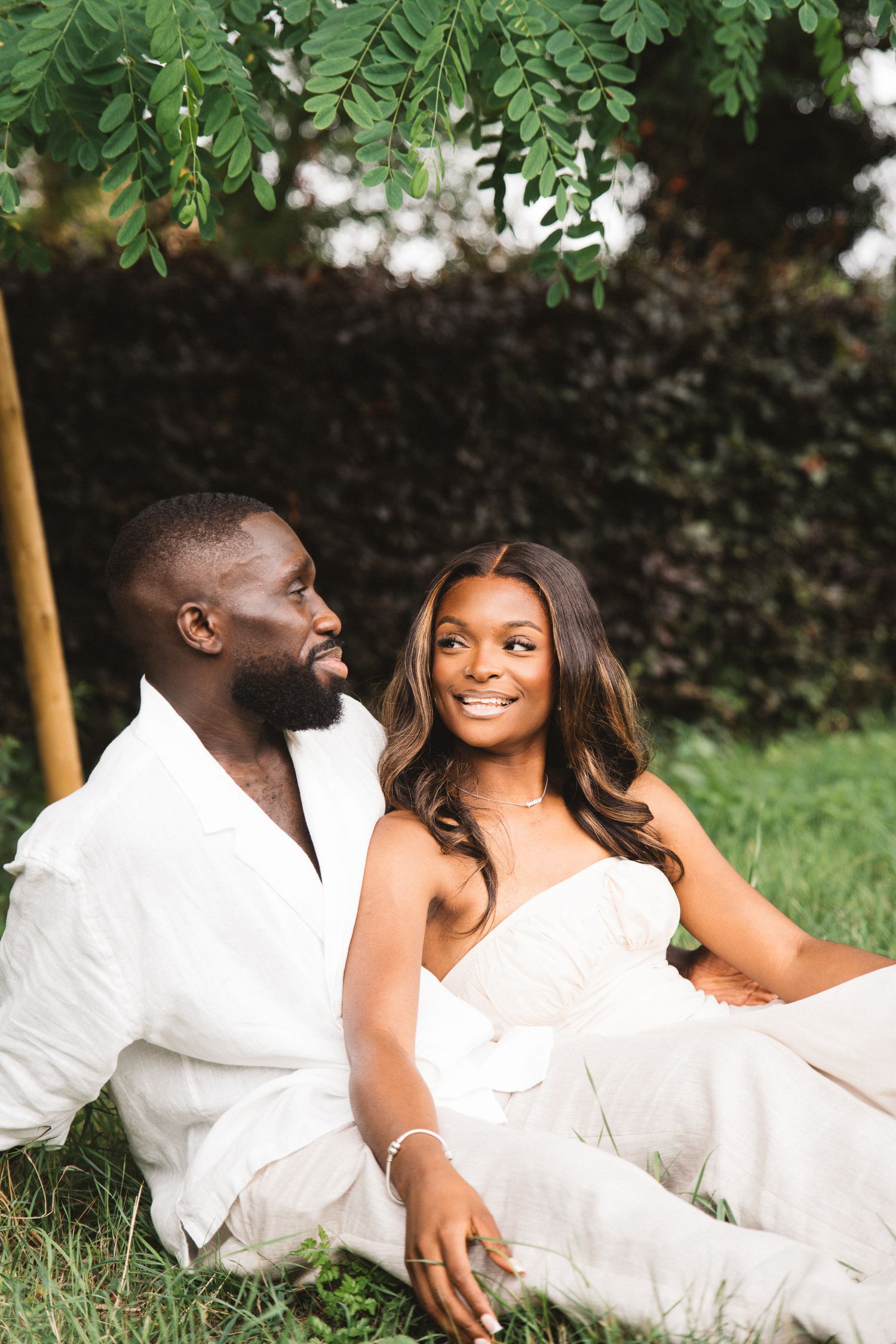 A couple in white outfits lying on grass under green foliage, sharing intimate moments.