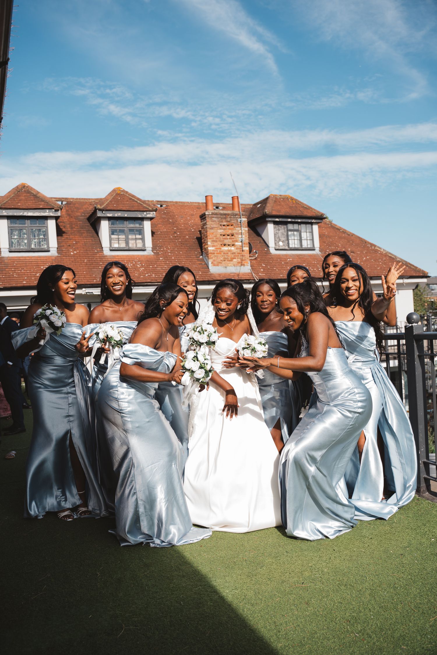 Bridesmaids in light blue dresses pose together on grass with a brick building backdrop.