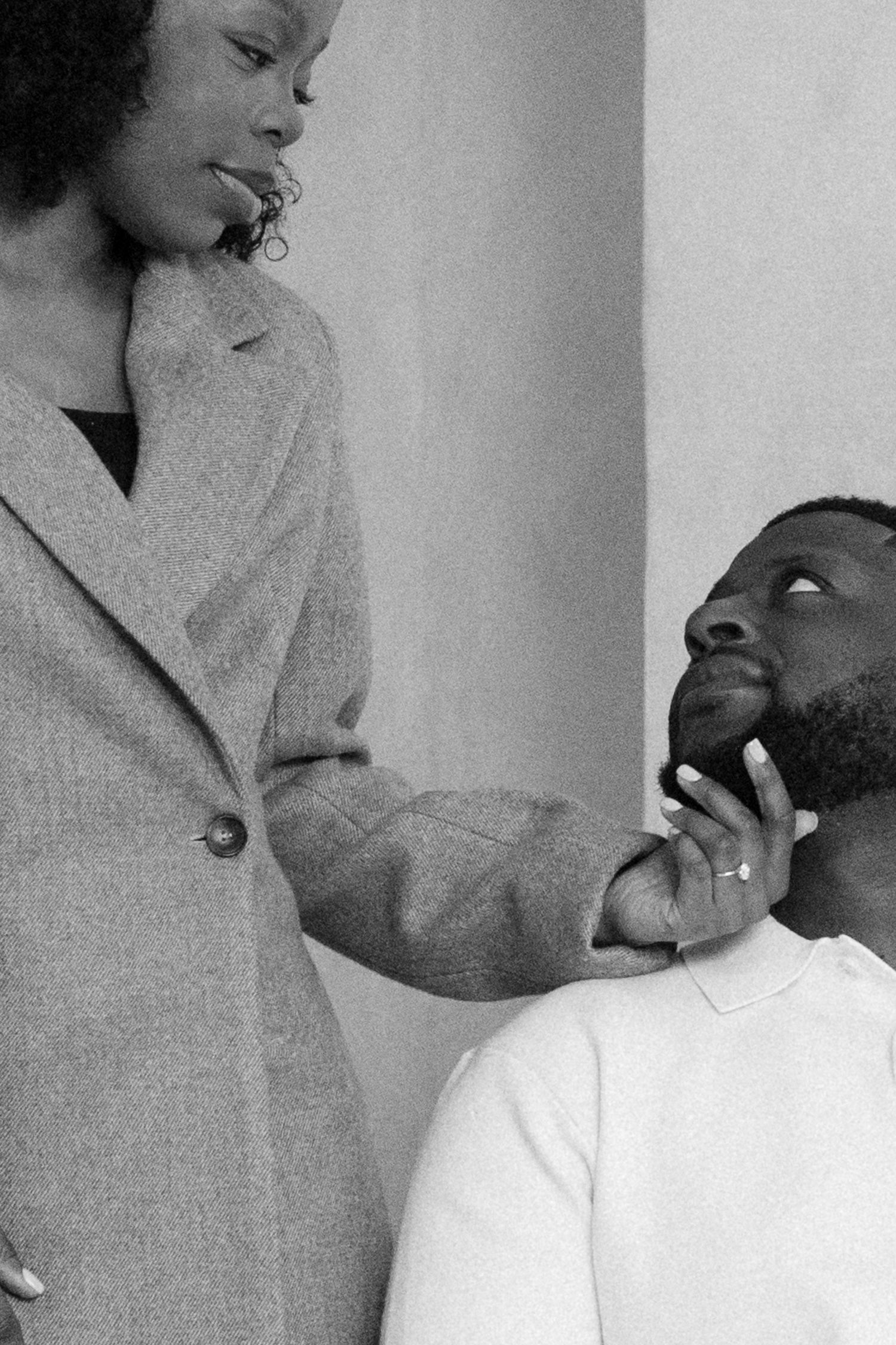 Black and white photo of a woman in a blazer tenderly holding a seated man's chin as they gaze at each other.