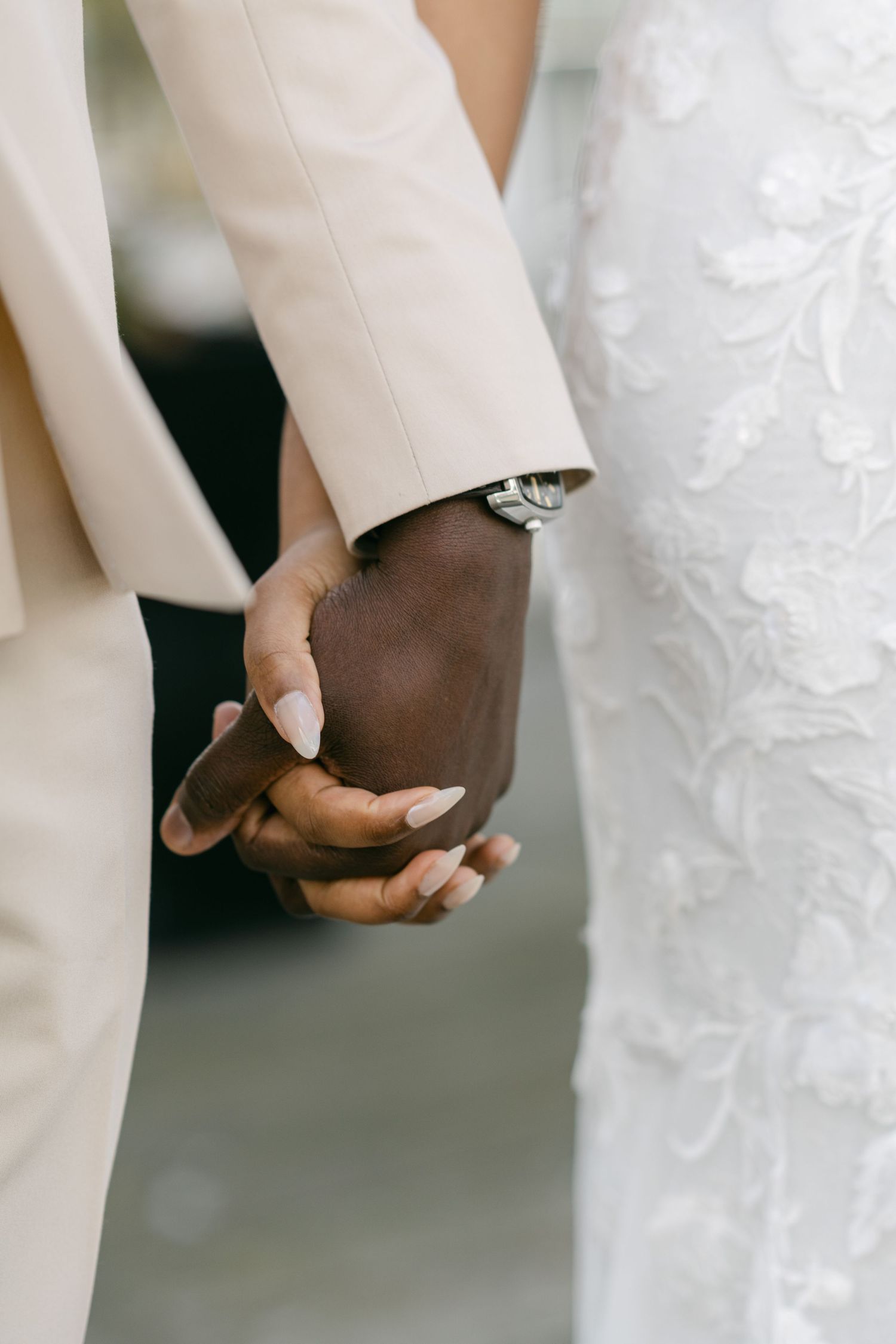 Close-up of newlyweds holding hands, showing white suit and lace dress details.