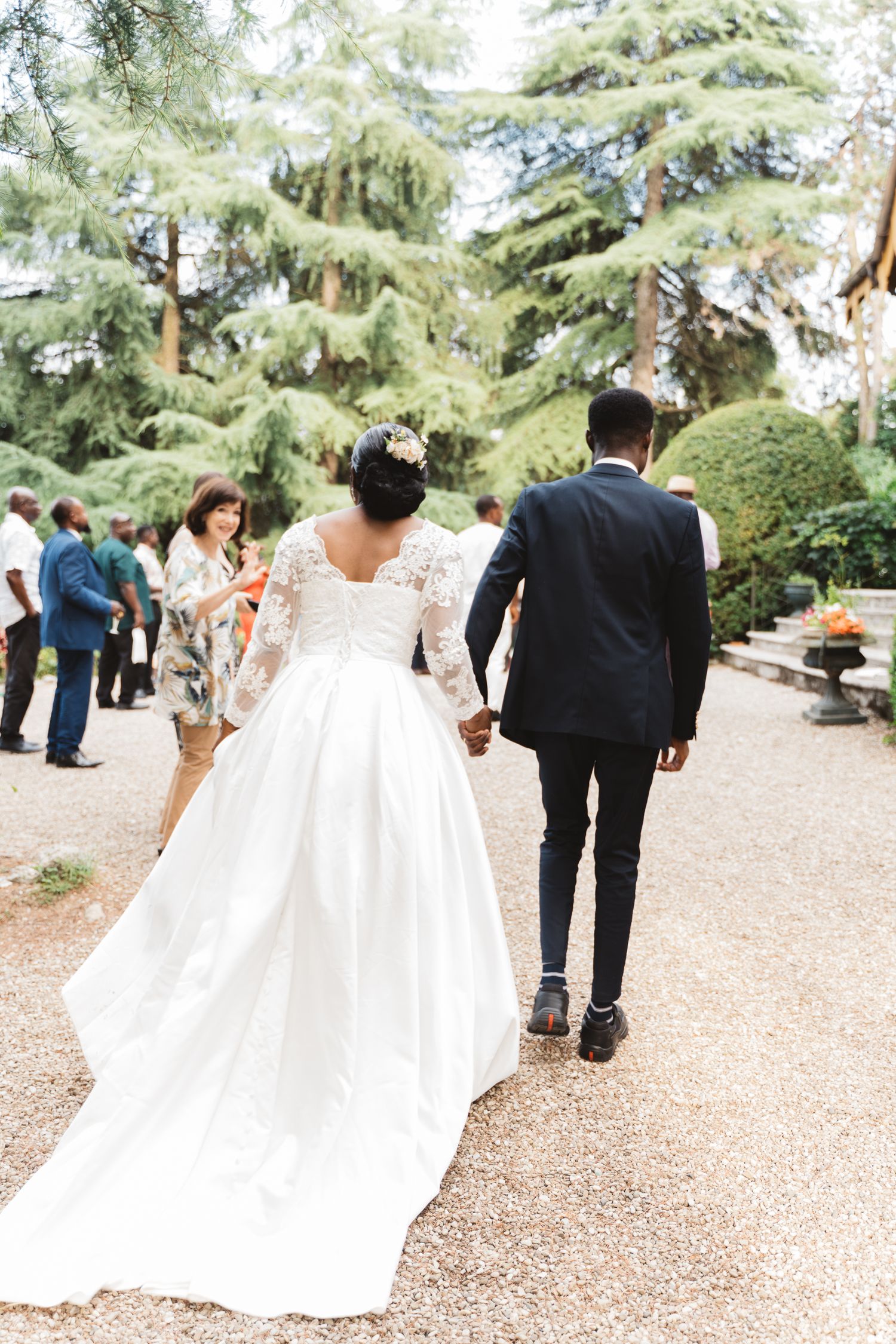 A flowing white wedding dress trails behind on a garden path during an outdoor celebration.