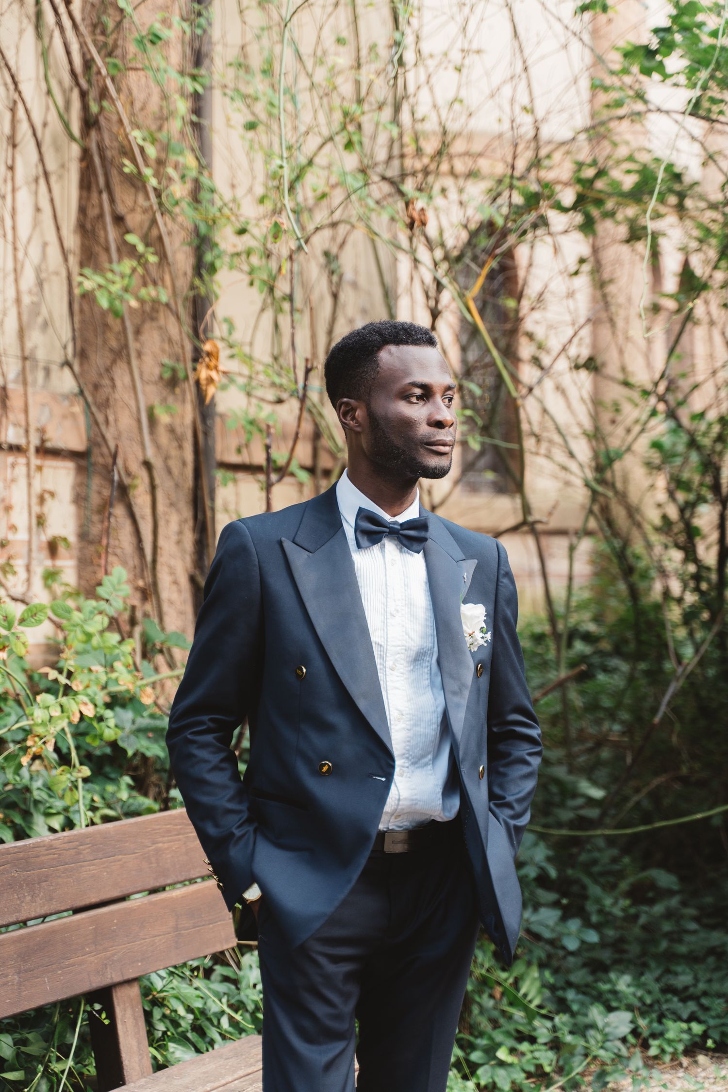 Groom in navy tuxedo posing on wooden bench in garden setting.
