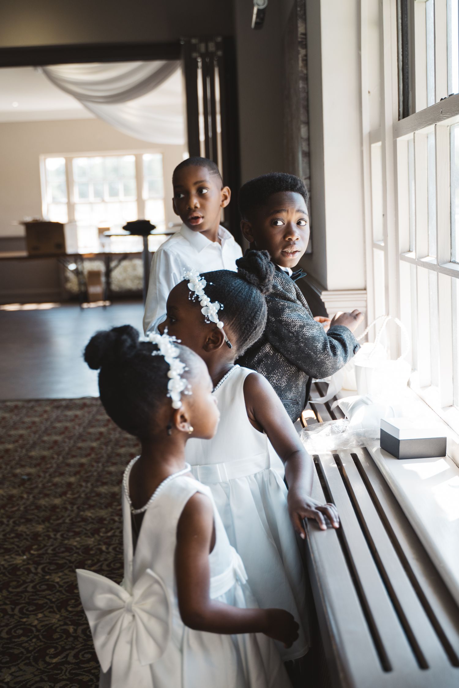 Young attendants in white dresses look out windows during wedding preparations.