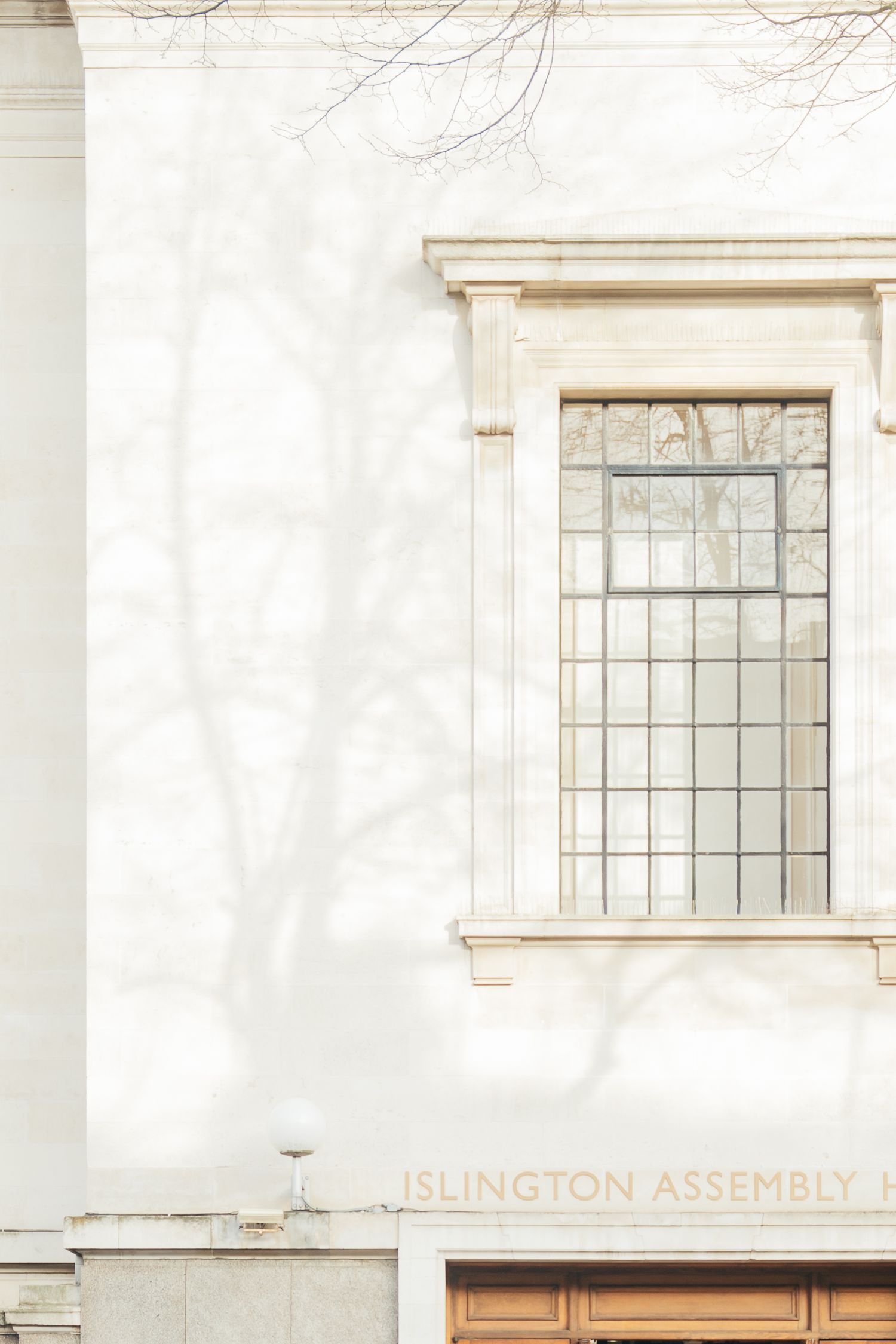 Islington Assembly Hall exterior with white stone facade, leaded glass window, and bare tree shadows.