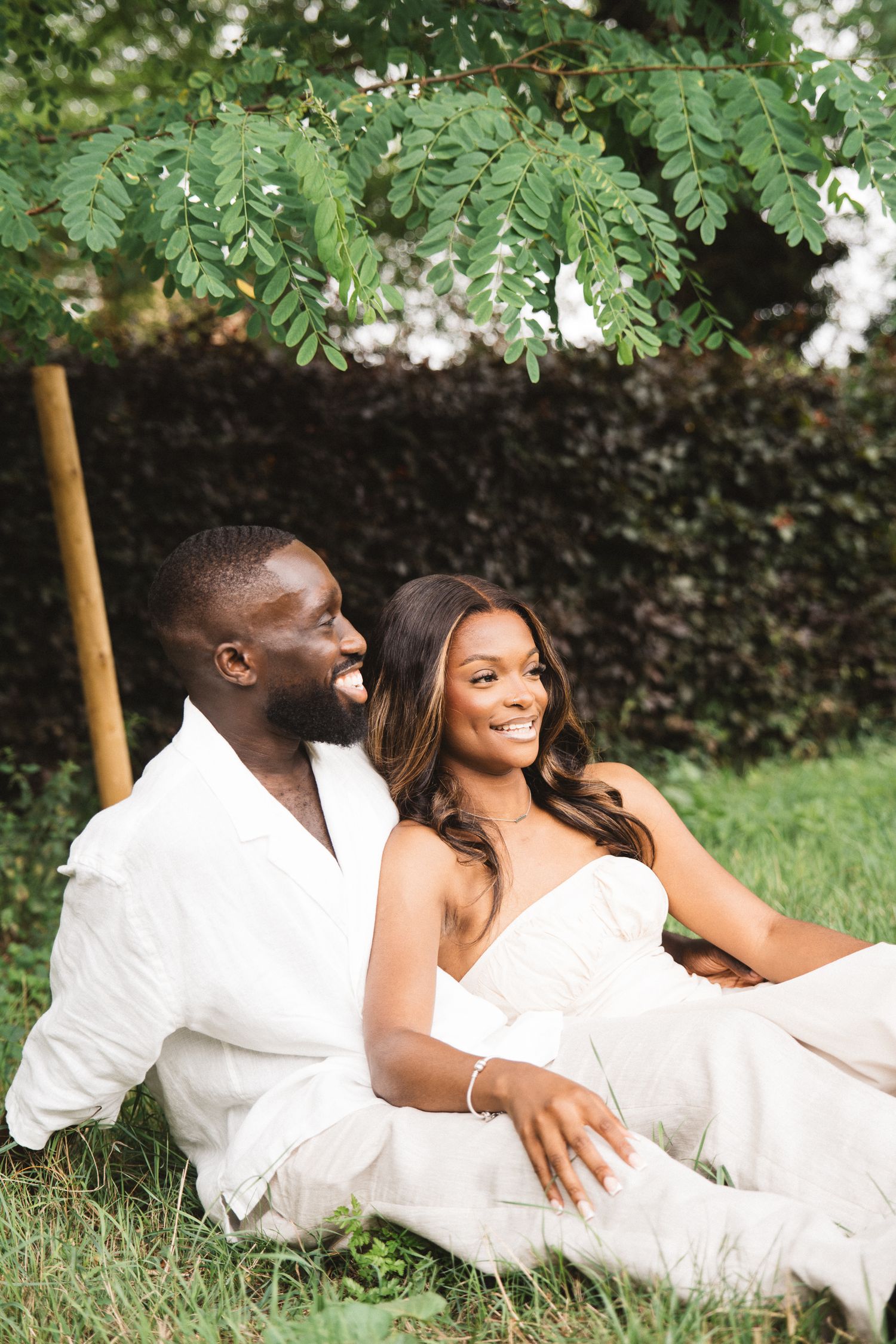 Romantic outdoor photoshoot with couple in white clothing relaxing on grass under trees.