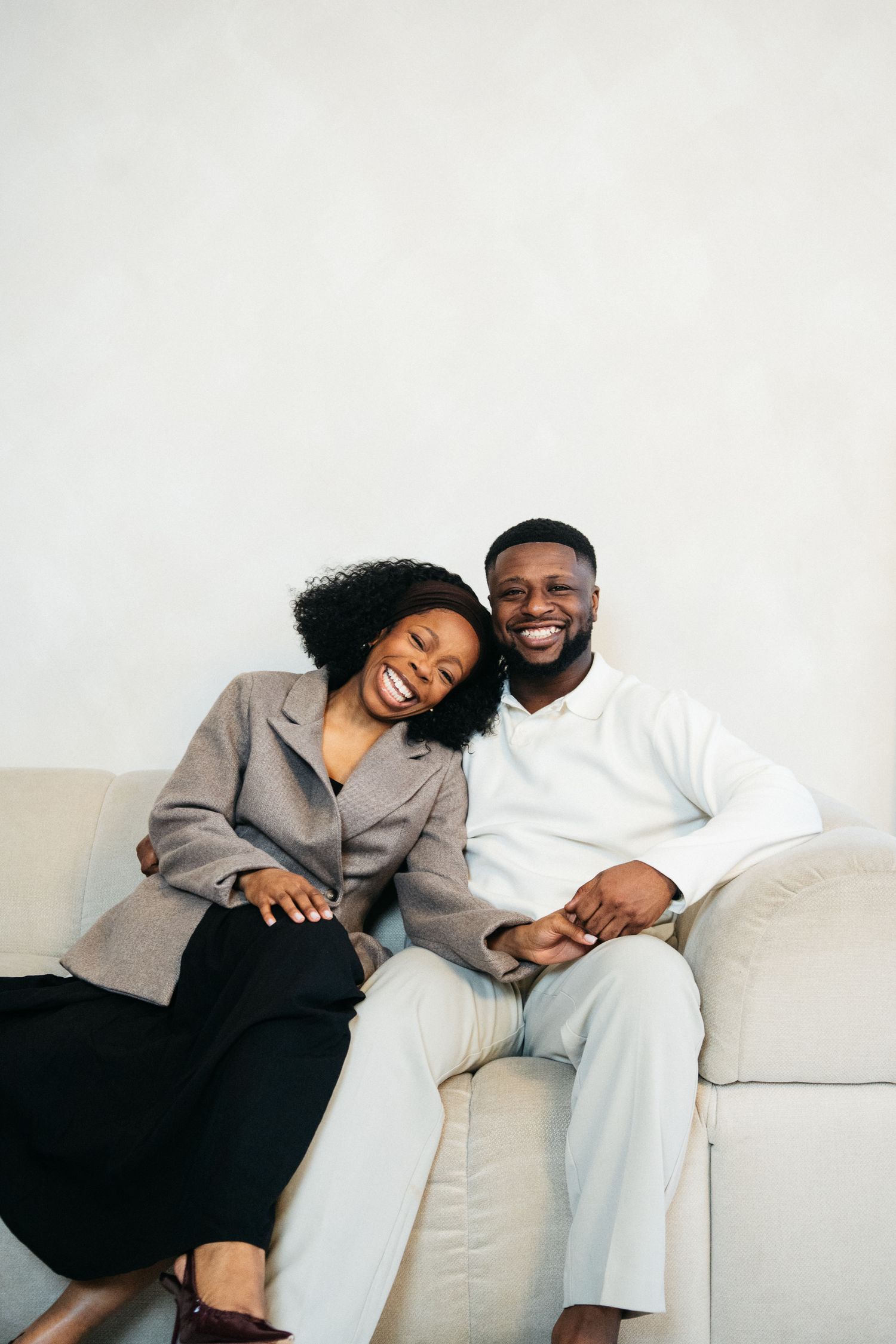 Happy Black couple sitting on a cream sofa, laughing and holding hands, woman in gray blazer leaning on mans shoulder.