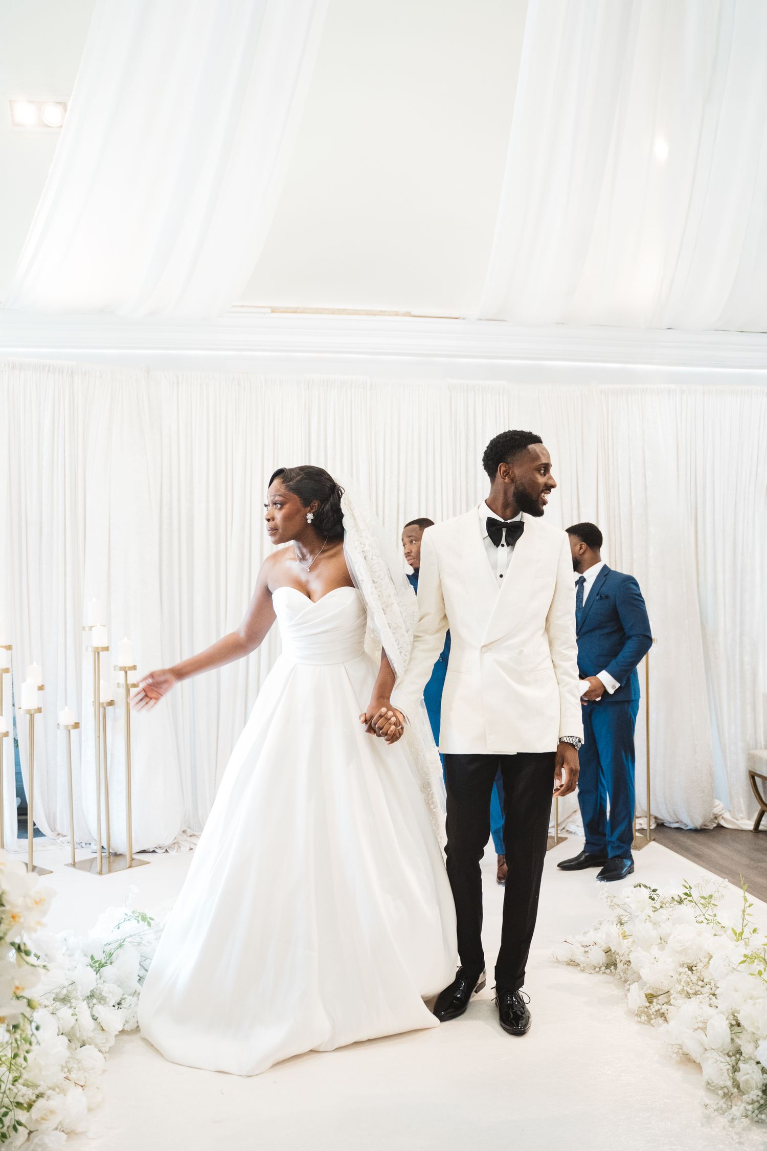 Wedding party in white and formal attire walking through an elegant draped venue.