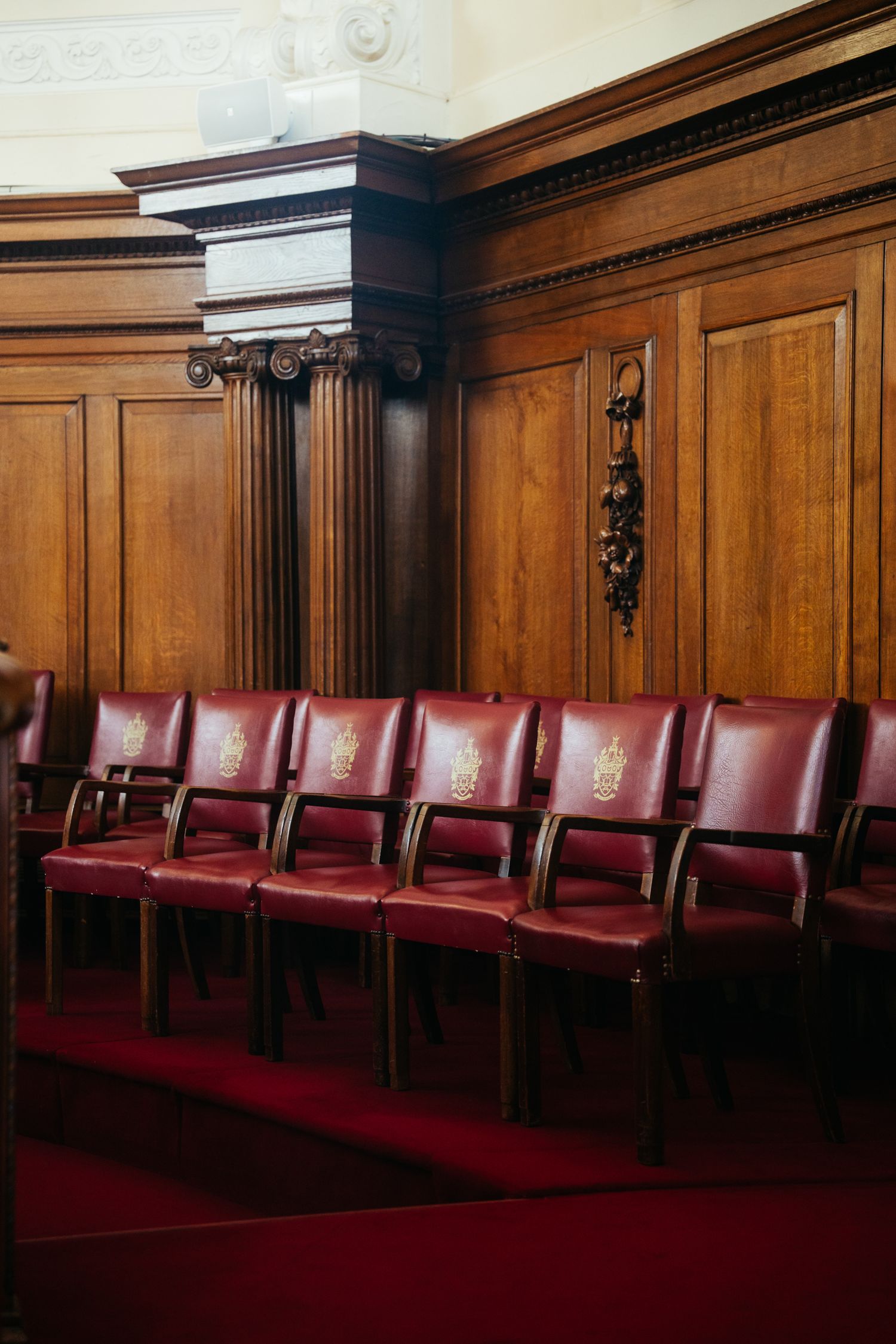 Row of vintage red leather chairs line a wood-paneled courtroom wall with ornate architectural details and molding.