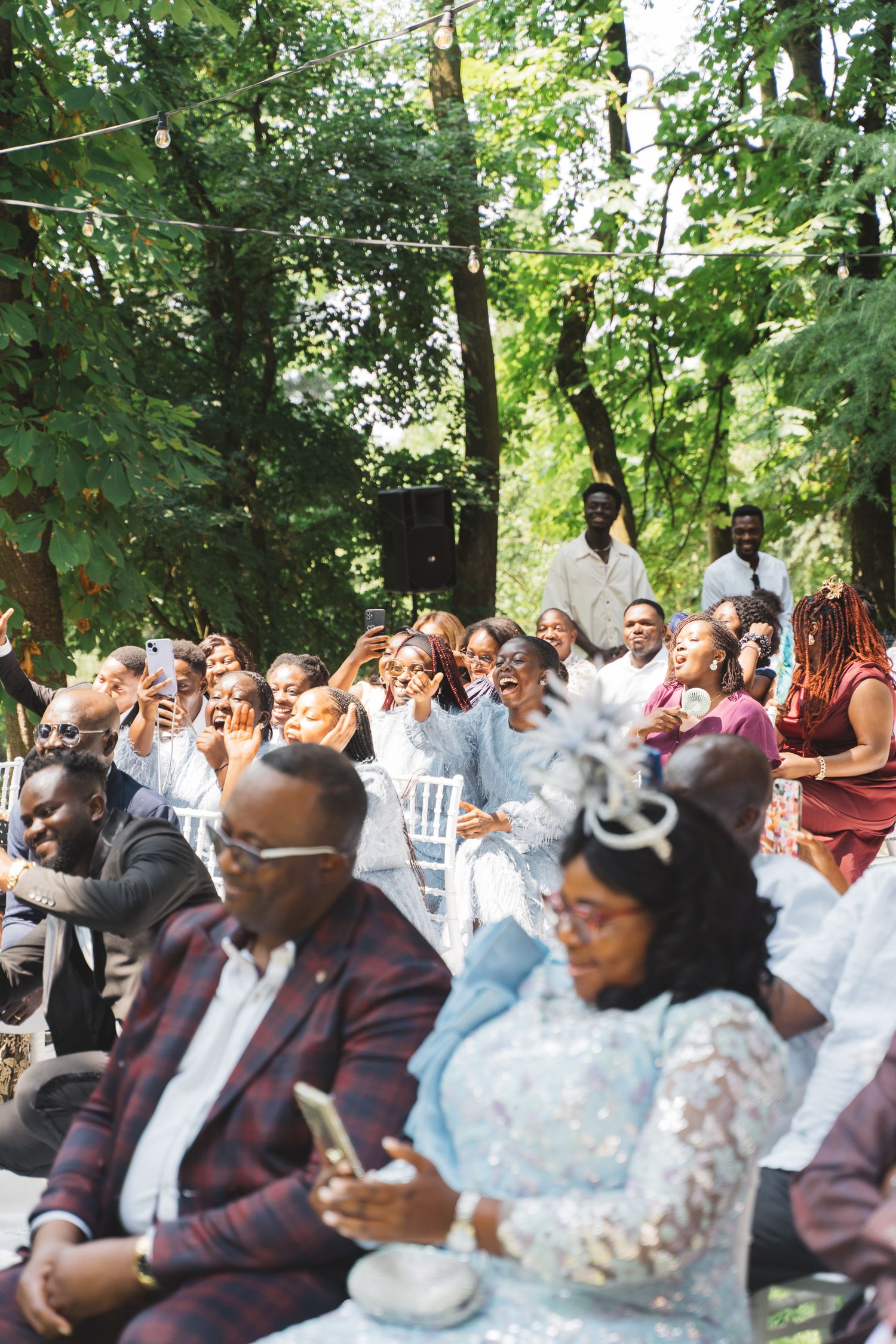 Wedding guests seated outdoors in a garden setting react with joy during a ceremony.