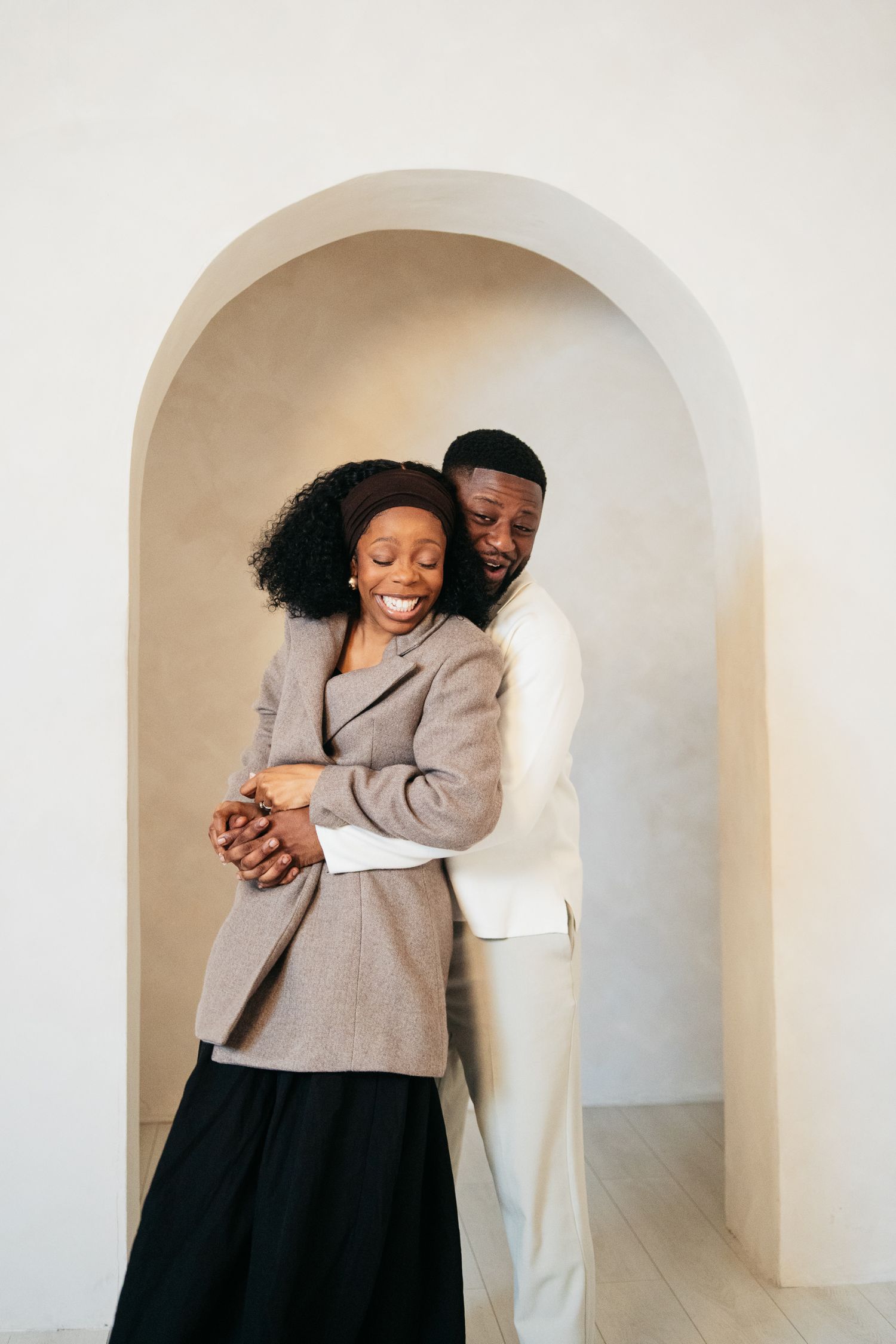 A joyful Black couple embraces warmly under an elegant arch, dressed in smart casual attire in a bright minimalist space.