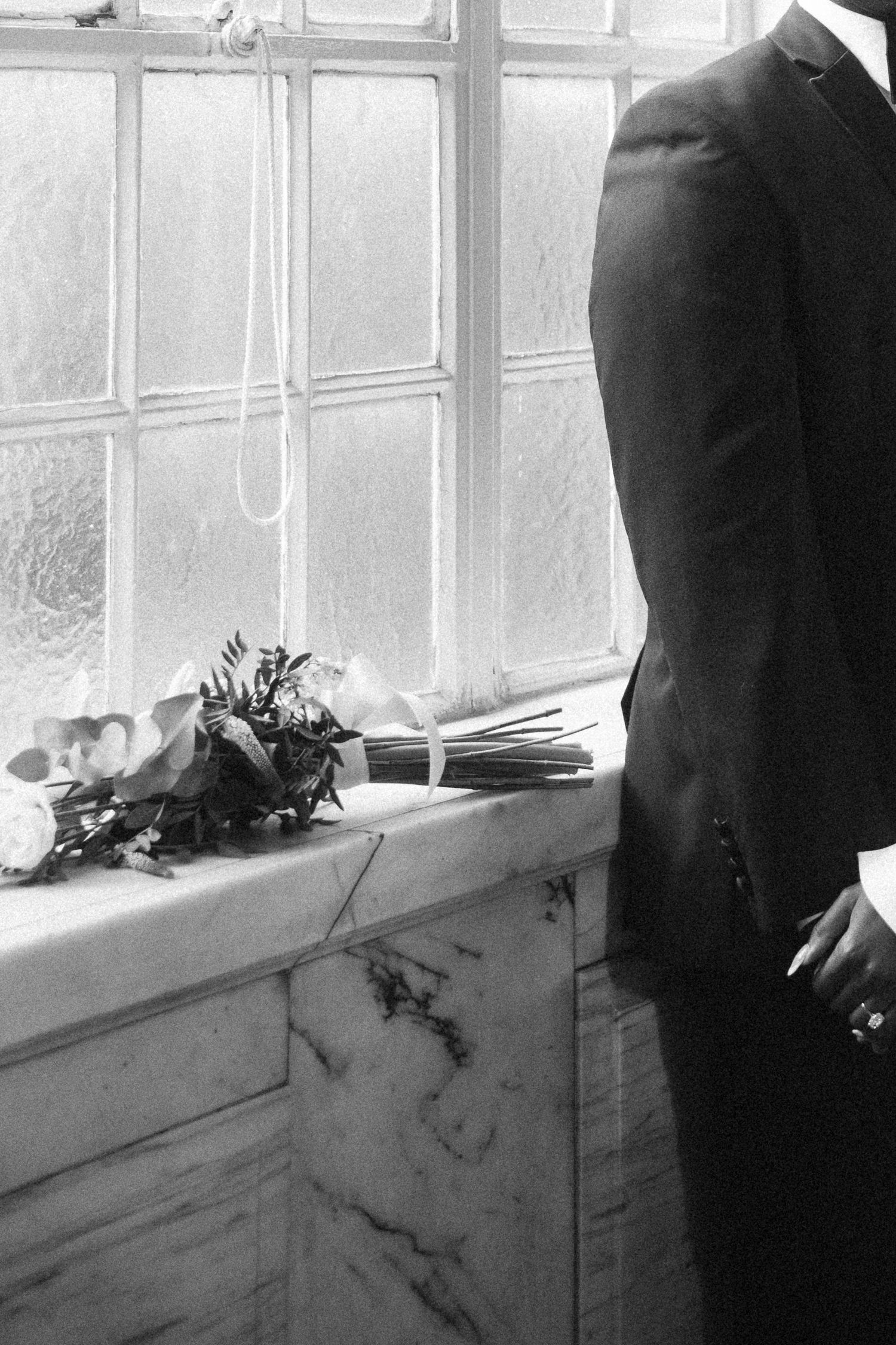 Black and white photo of a groom in a suit standing by a marble windowsill with a bridal bouquet.