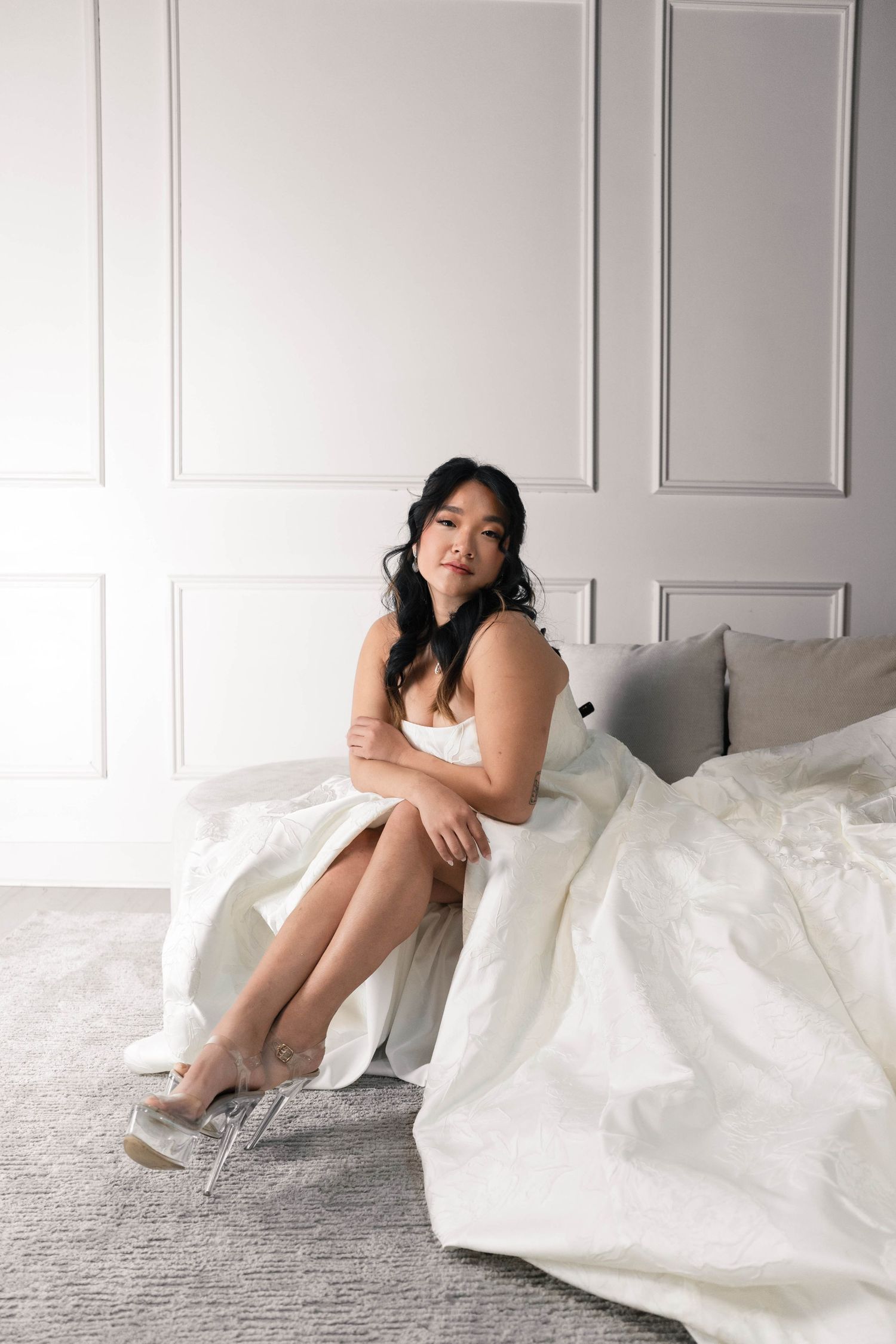 Bride in white wedding dress sitting on bed's edge wearing clear heels in elegant white paneled room.