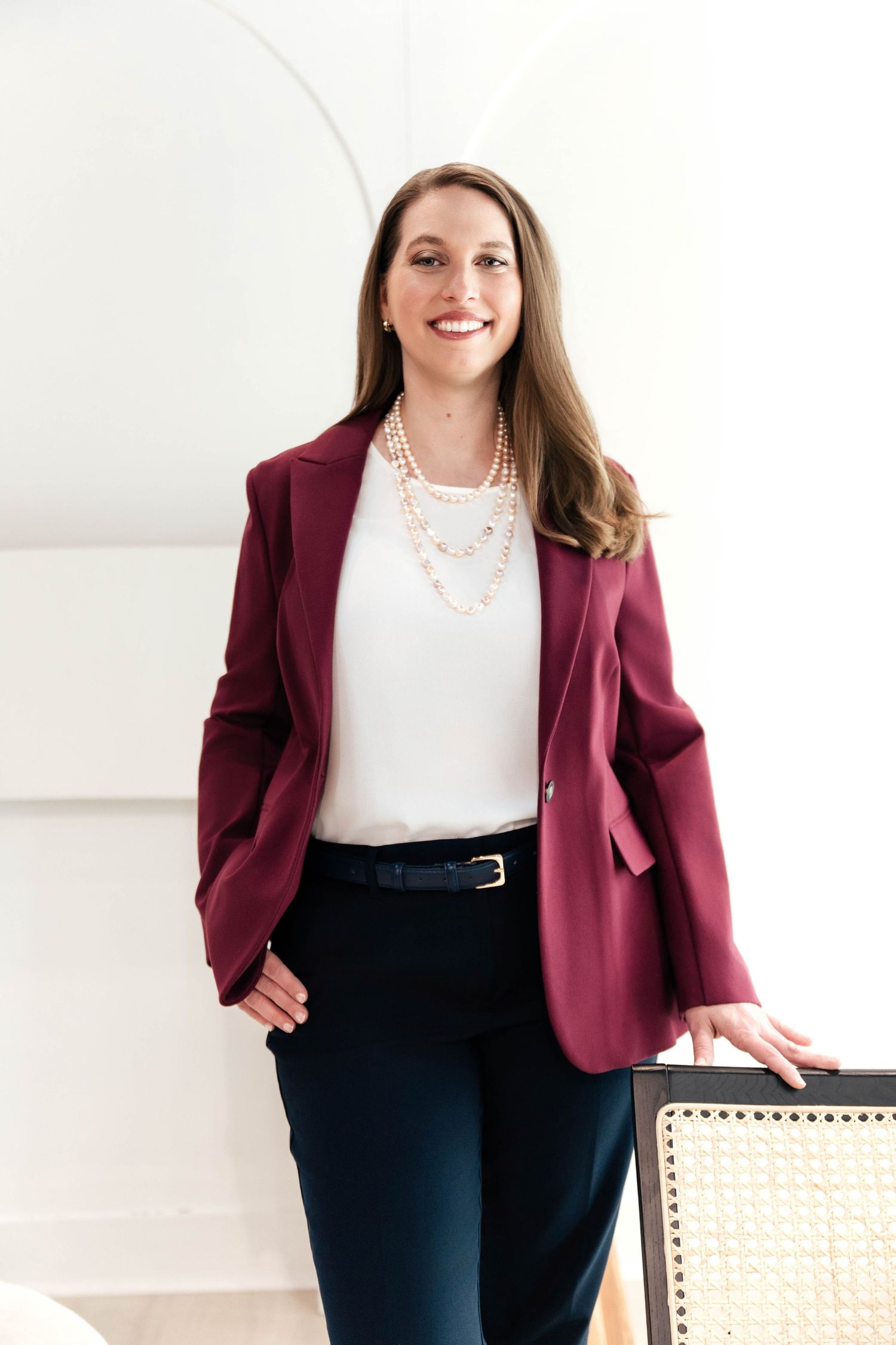 Professional woman in burgundy blazer and pearl necklace smiling confidently in a bright modern office setting.