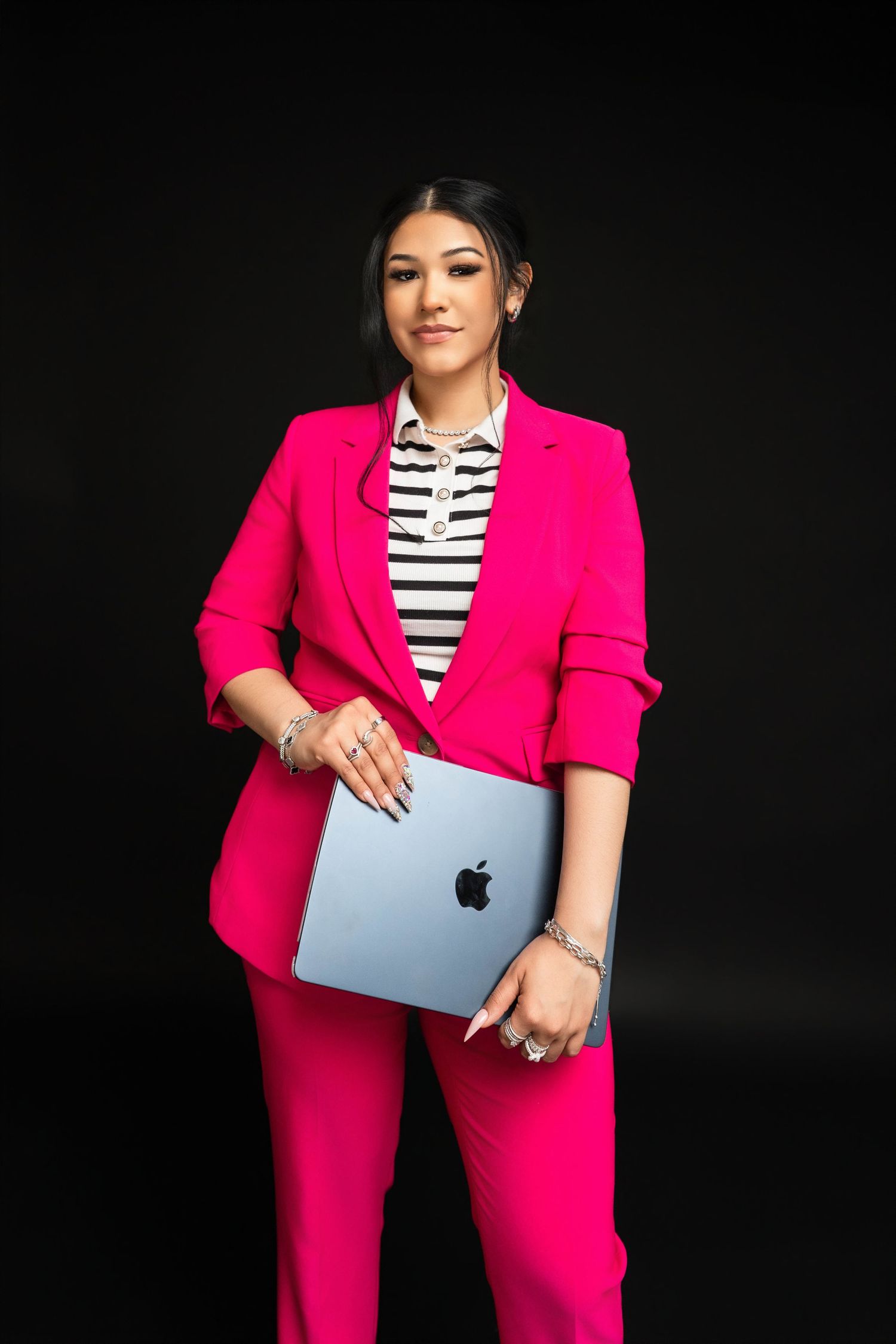 Woman in pink blazer and pants holds MacBook, wearing striped shirt, posing confidently against black background.