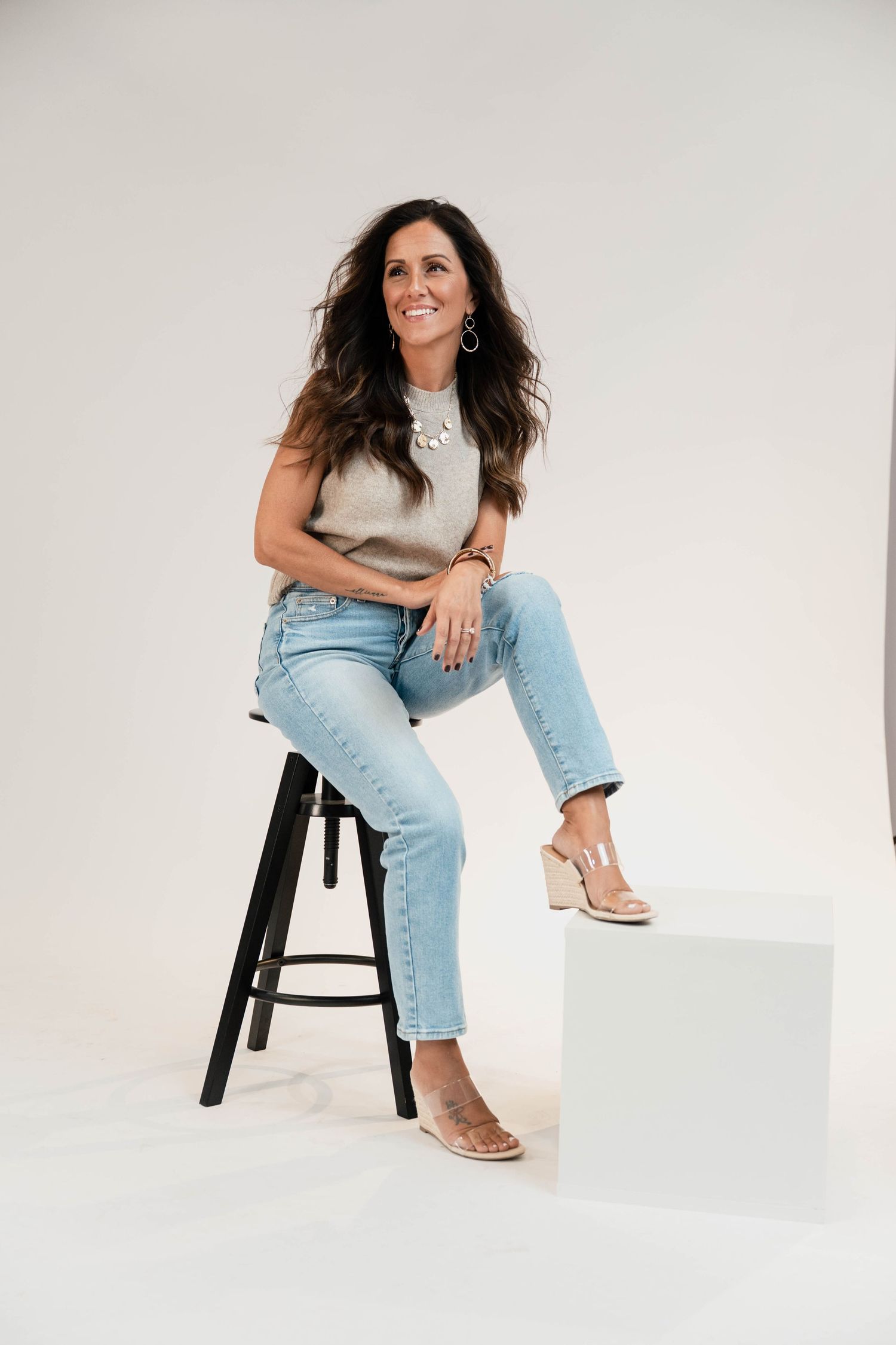 Woman in light jeans and gray top sitting on stool, wearing wedge sandals and silver necklace, smiling in studio.