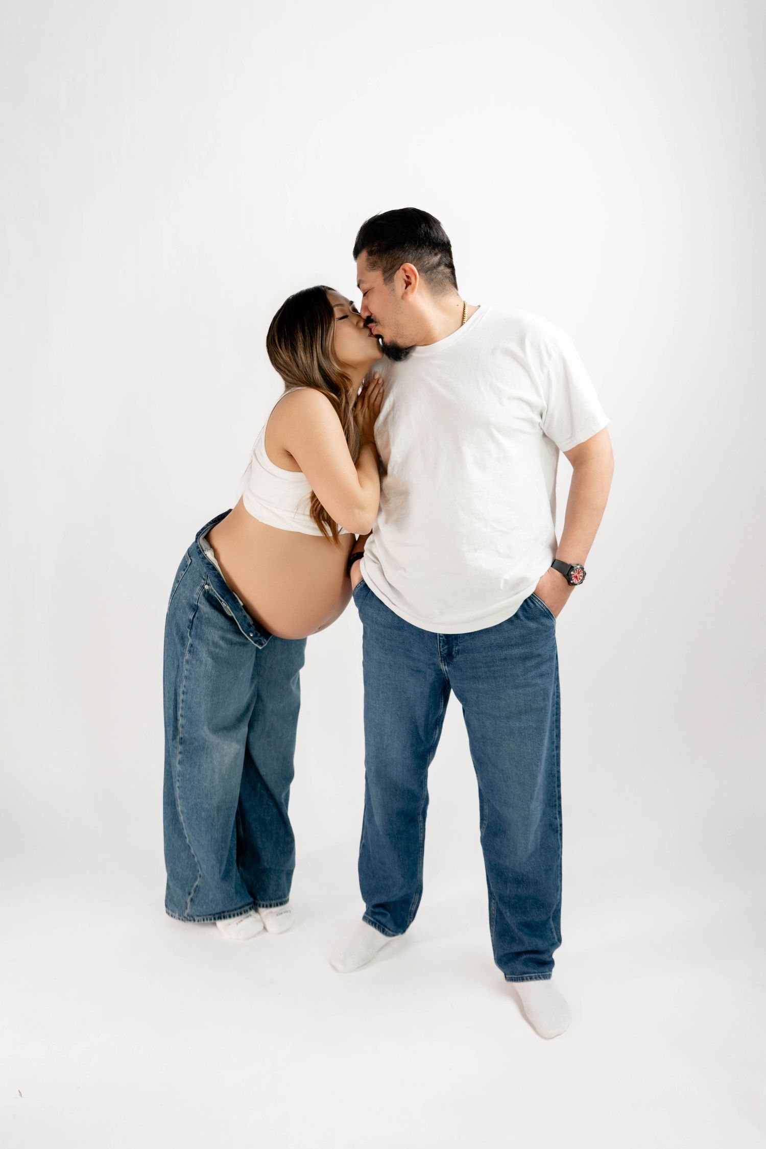 Pregnant couple kissing in studio, woman in white crop top and jeans showing baby bump, man in white t-shirt and jeans.
