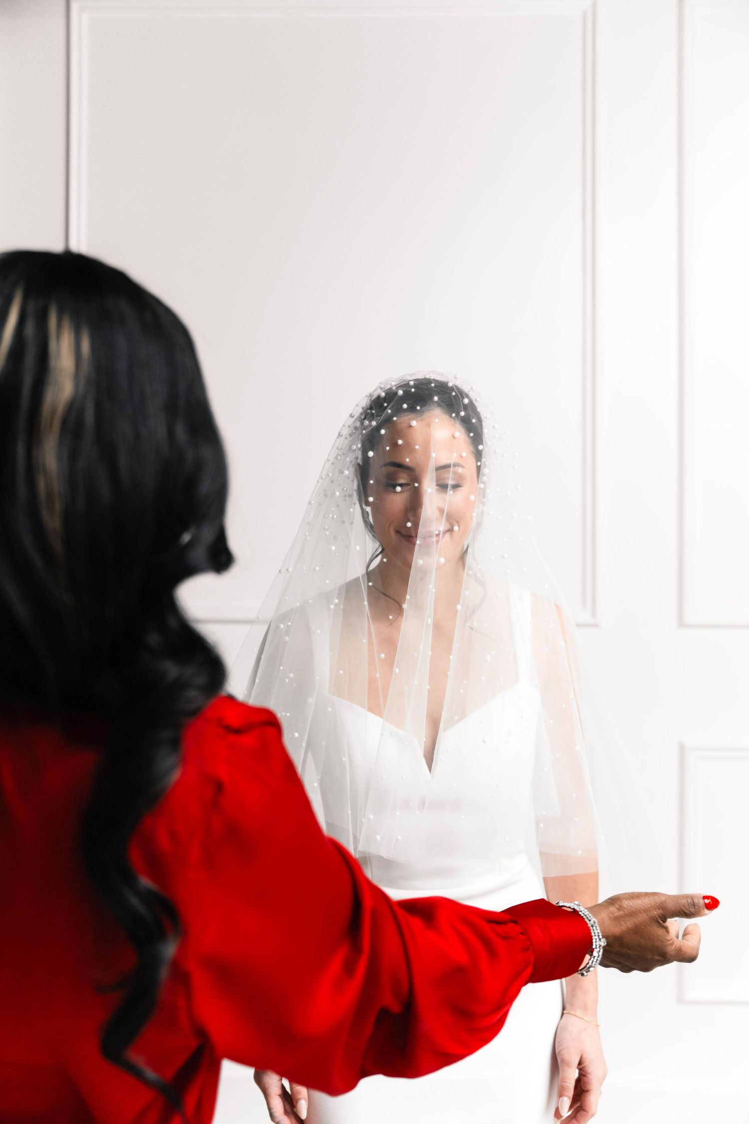A smiling bride in a white gown and pearl veil stands before a woman in a red dress during wedding preparations.
