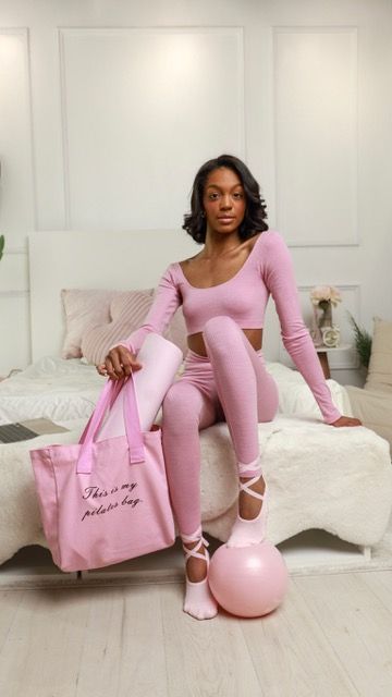 Woman in pink activewear set holding a pink tote bag beside a pilates ball on a white bed.
