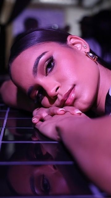 Young woman with glam makeup rests her chin on hands, reflecting on a mirrored surface under purple lighting.
