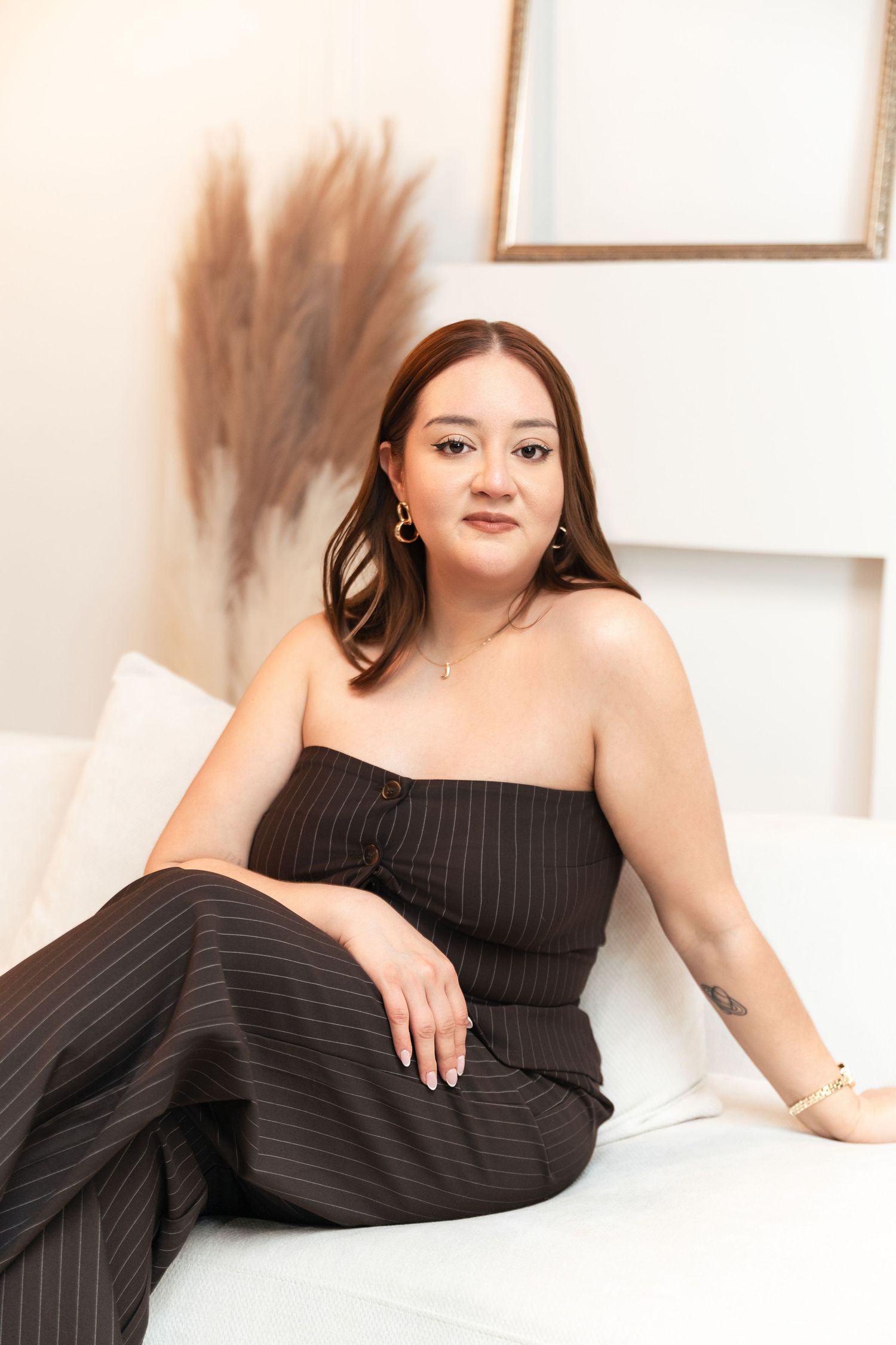Woman in black pinstripe strapless top and wide-leg pants sitting on white sofa with dried pampas grass decor.