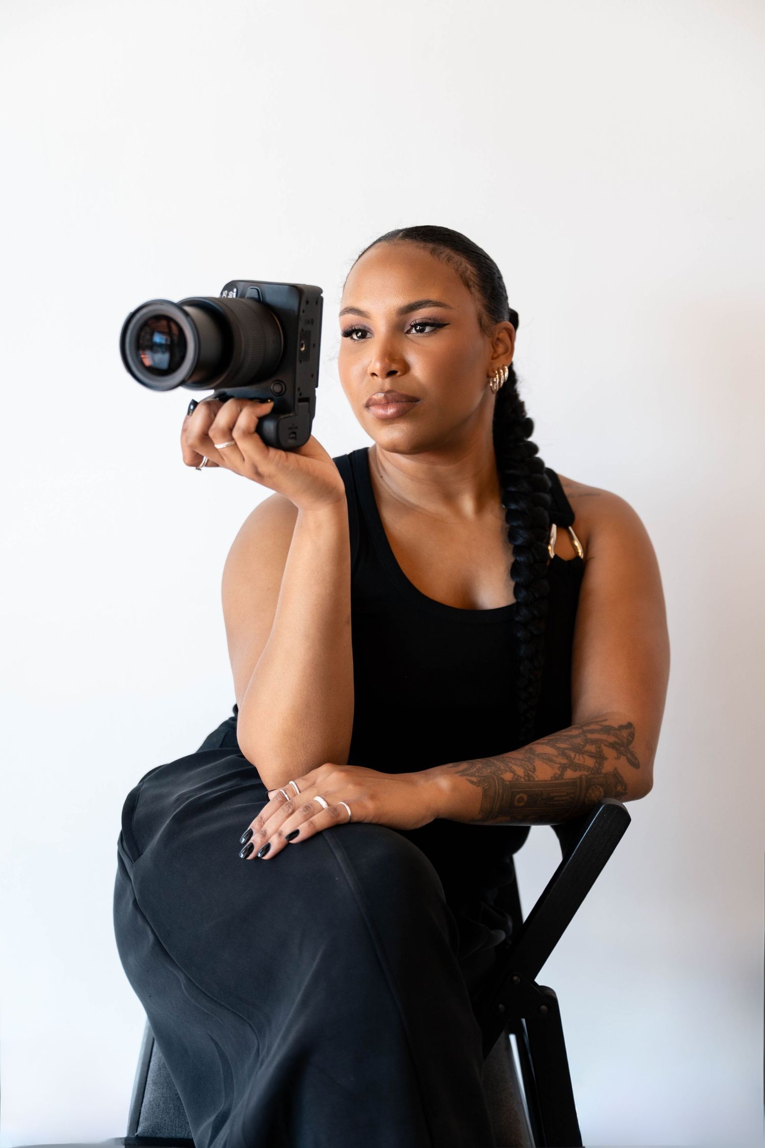 A woman in black attire sits holding a professional camera, showcasing tattoos and braided hair against a white background.