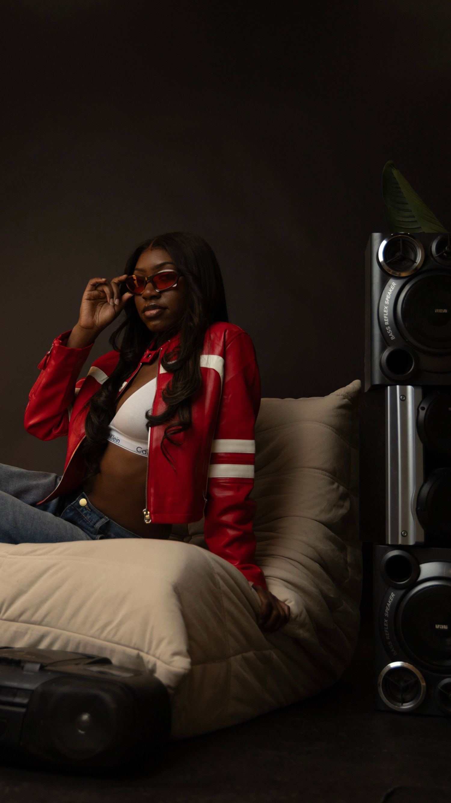 A young woman in a red leather jacket and sunglasses poses on a bean bag chair next to a stereo speaker.