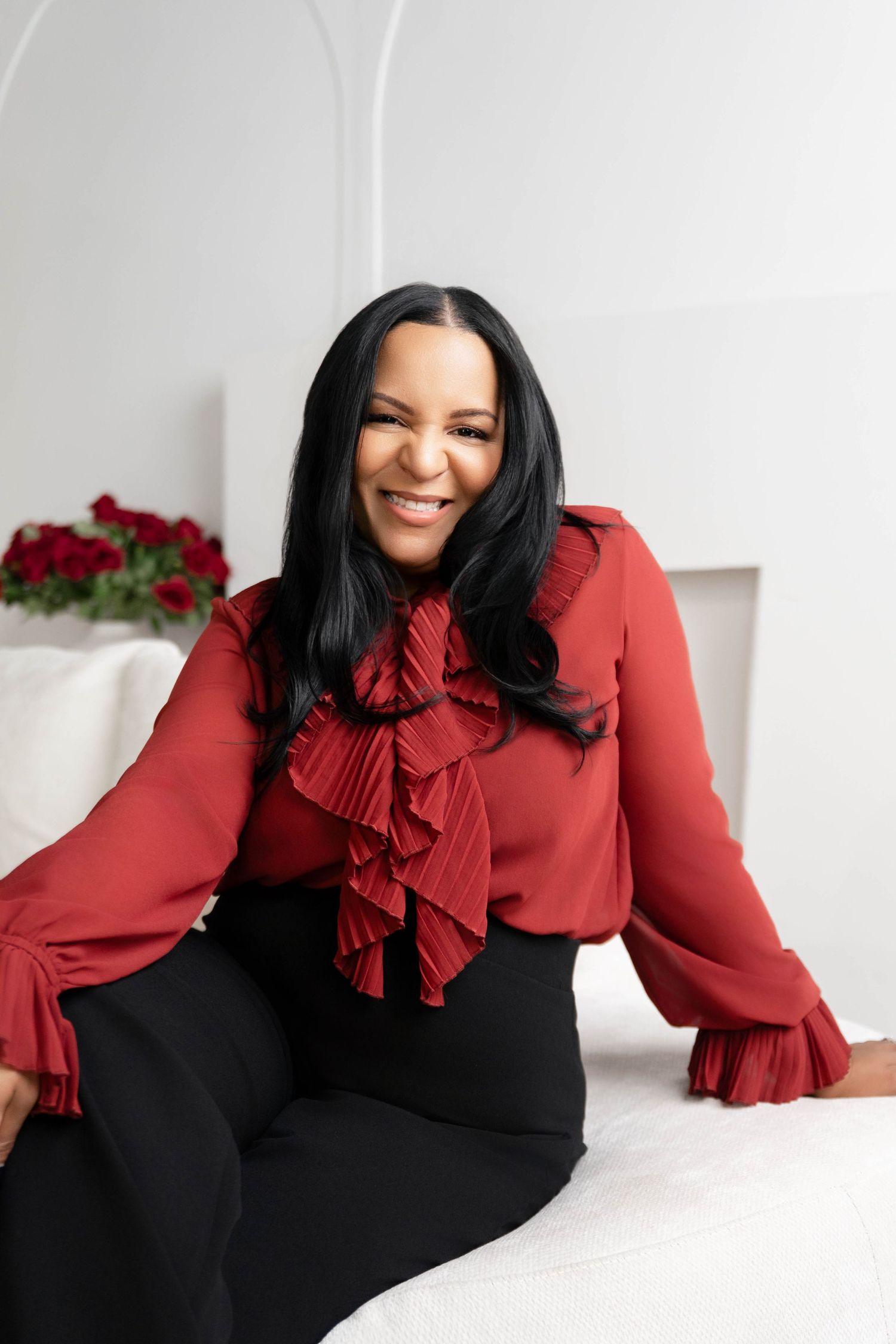 Smiling woman in red ruffled blouse and black pants sitting on white surface with roses in background.
