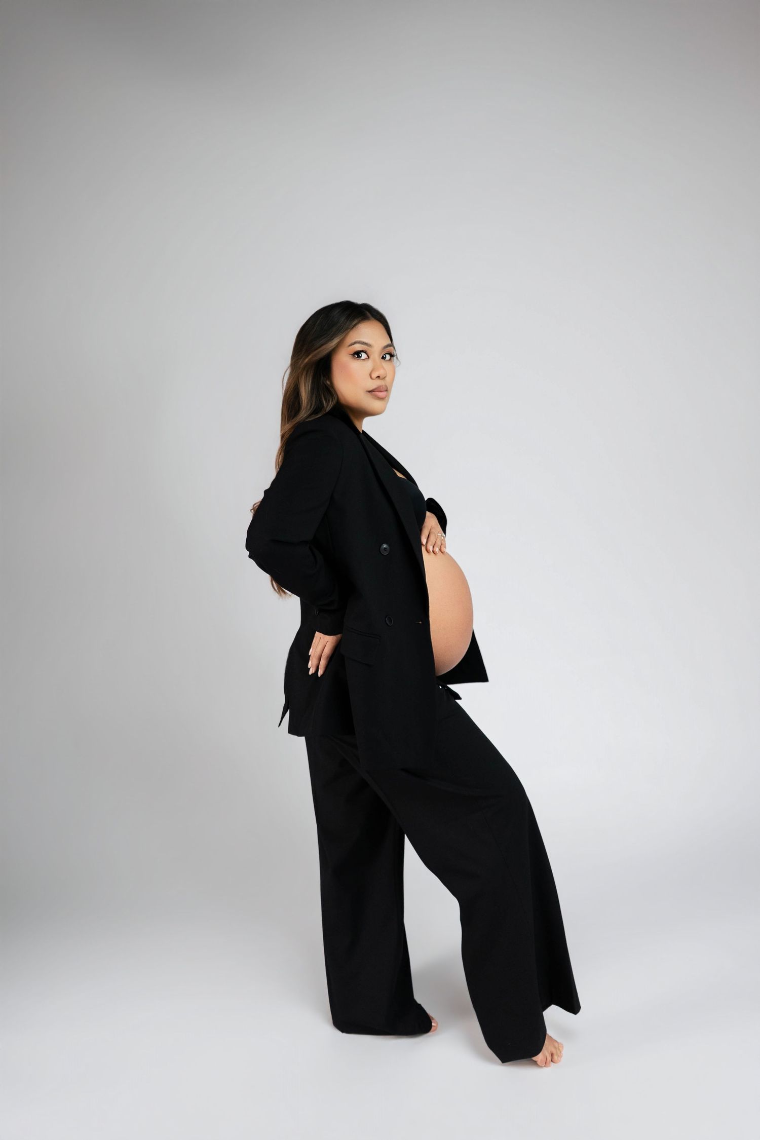Pregnant woman in black blazer and wide-leg pants posing sideways, cradling baby bump in a studio setting.