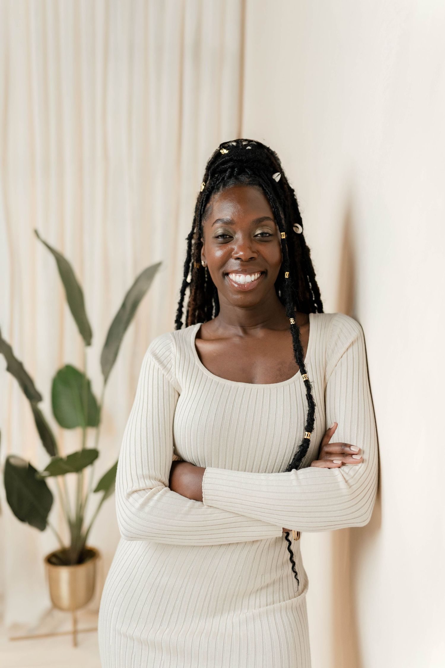Smiling Black woman with braids, arms crossed, wearing a cream ribbed dress, standing against a light wall with a plant.