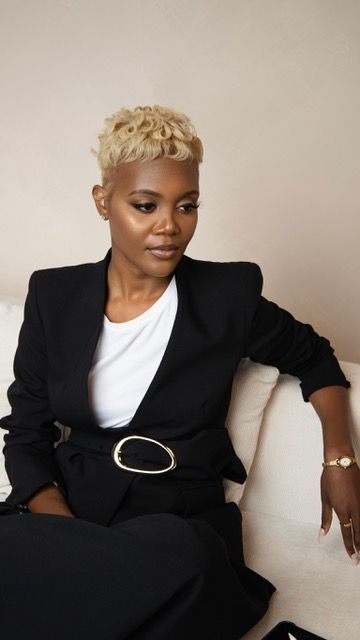 Black woman with short blonde curly hair wearing a black blazer and white tee, seated on a white sofa.